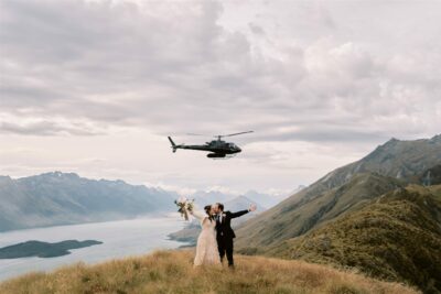 queenstown new zealand heli wedding elopement photographer videographer | A newlywed couple celebrates their Queenstown elopement on a mountain ridge with a helicopter flying overhead against a backdrop of a vast lake and rolling hills.