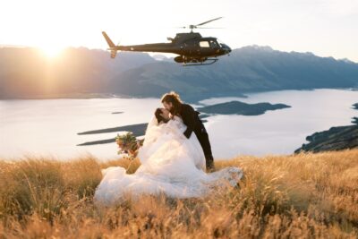 queenstown new zealand heli wedding elopement photographer videographer | A couple in wedding attire kisses on a grassy hill at sunset during their Queenstown elopement, with a helicopter hovering above and a scenic lake and mountain range in the background—a perfect moment captured by masterful wedding photography.