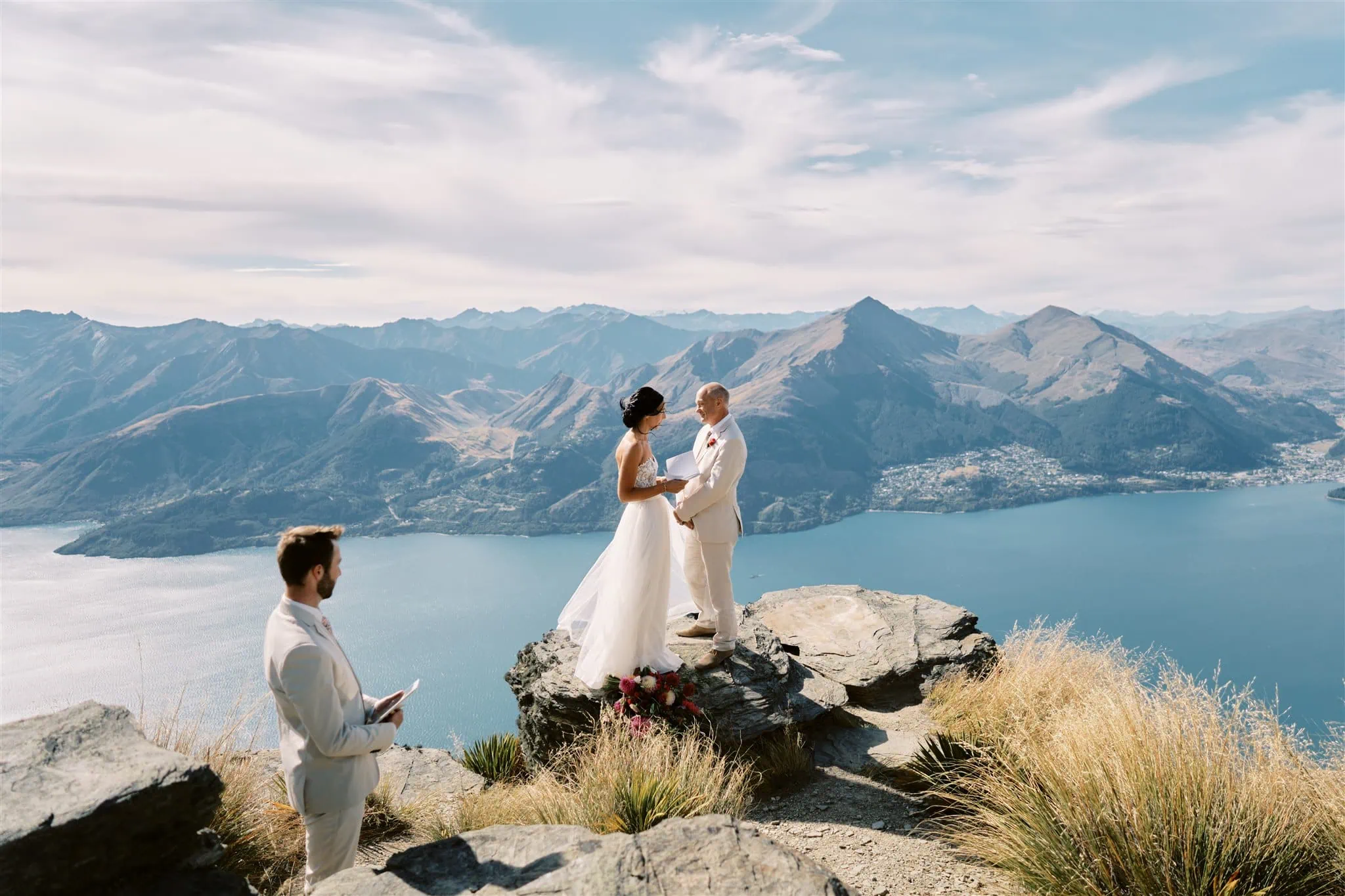 queenstown new zealand heli wedding elopement photographer videographer | A wedding ceremony on a mountaintop in Queenstown with a couple standing hand-in-hand, a guitarist seated nearby, and expansive views of a lake and mountains in the background.