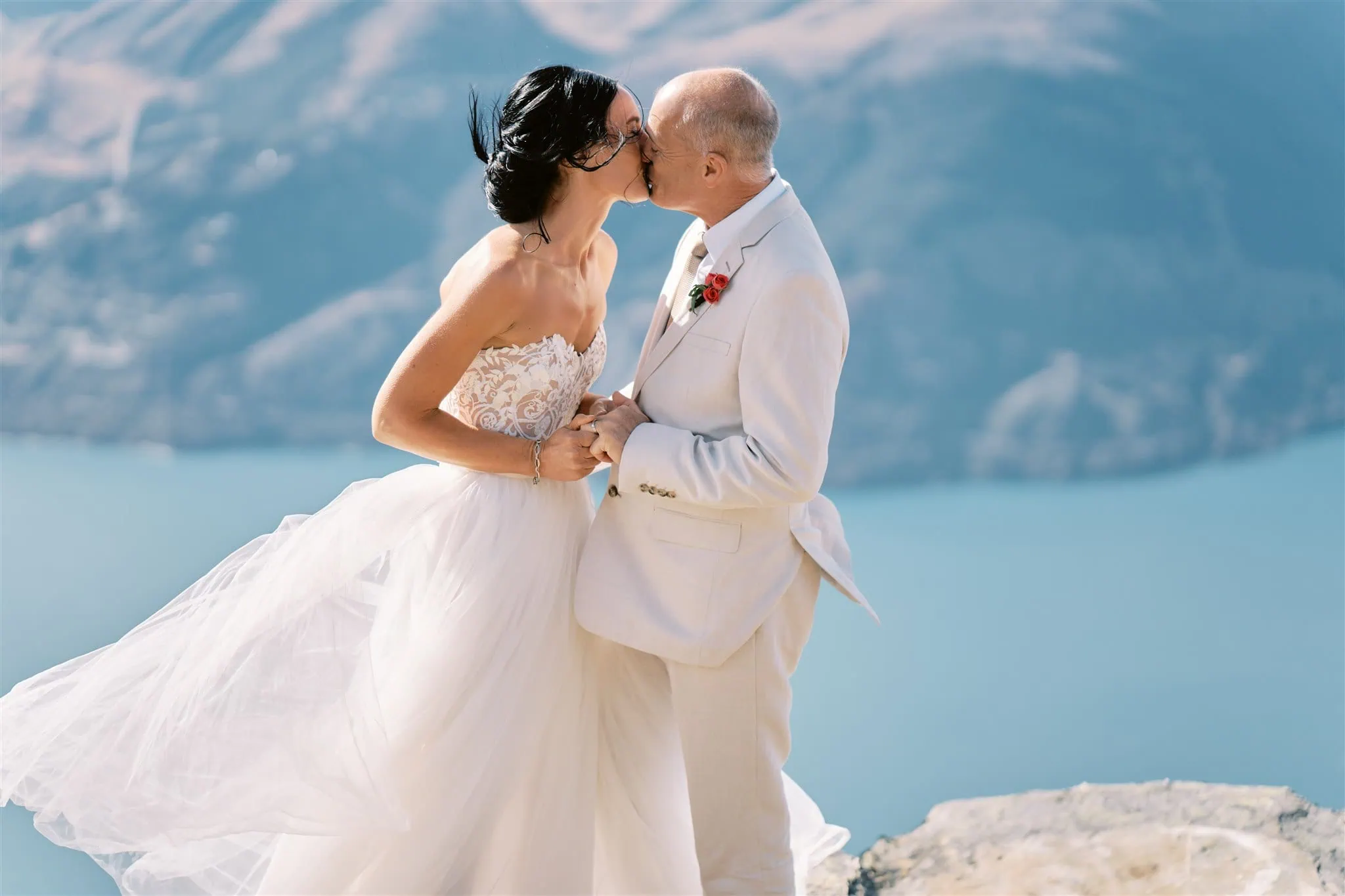 queenstown new zealand heli wedding elopement photographer videographer | Ian & Vivienne, a bride and groom, kissing on a cliff with a serene blue lake and mountains in the background during their Queenstown elopement.
