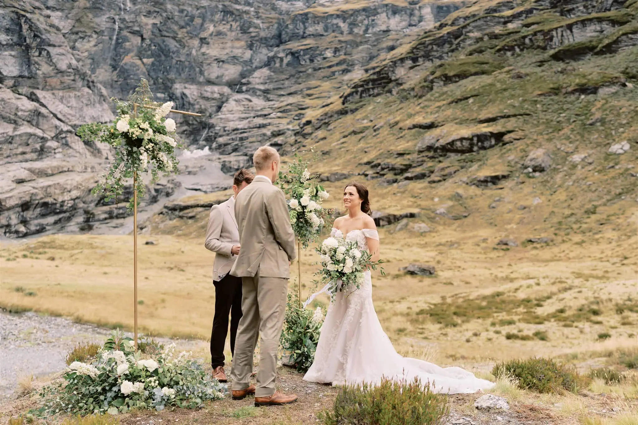 queenstown new zealand heli wedding elopement photographer videographer | A wedding ceremony taking place outdoors in Queenstown with a couple standing under a floral arch, surrounded by a rugged mountainous backdrop.