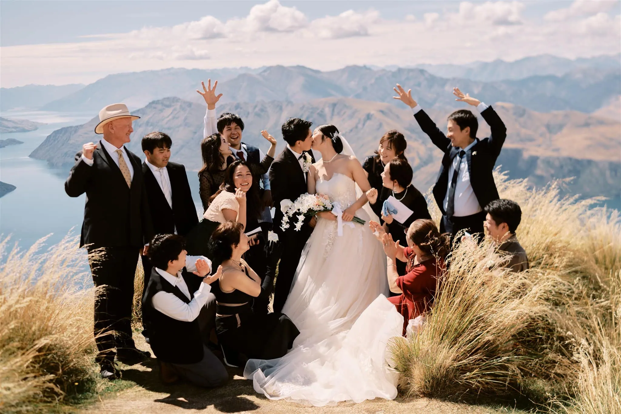 queenstown new zealand heli wedding elopement photographer videographer | A joyful Queenstown elopement group photo on a mountain overlook with Ryo & Tamano kissing and guests cheering around them, scenic lake and mountains in the background.