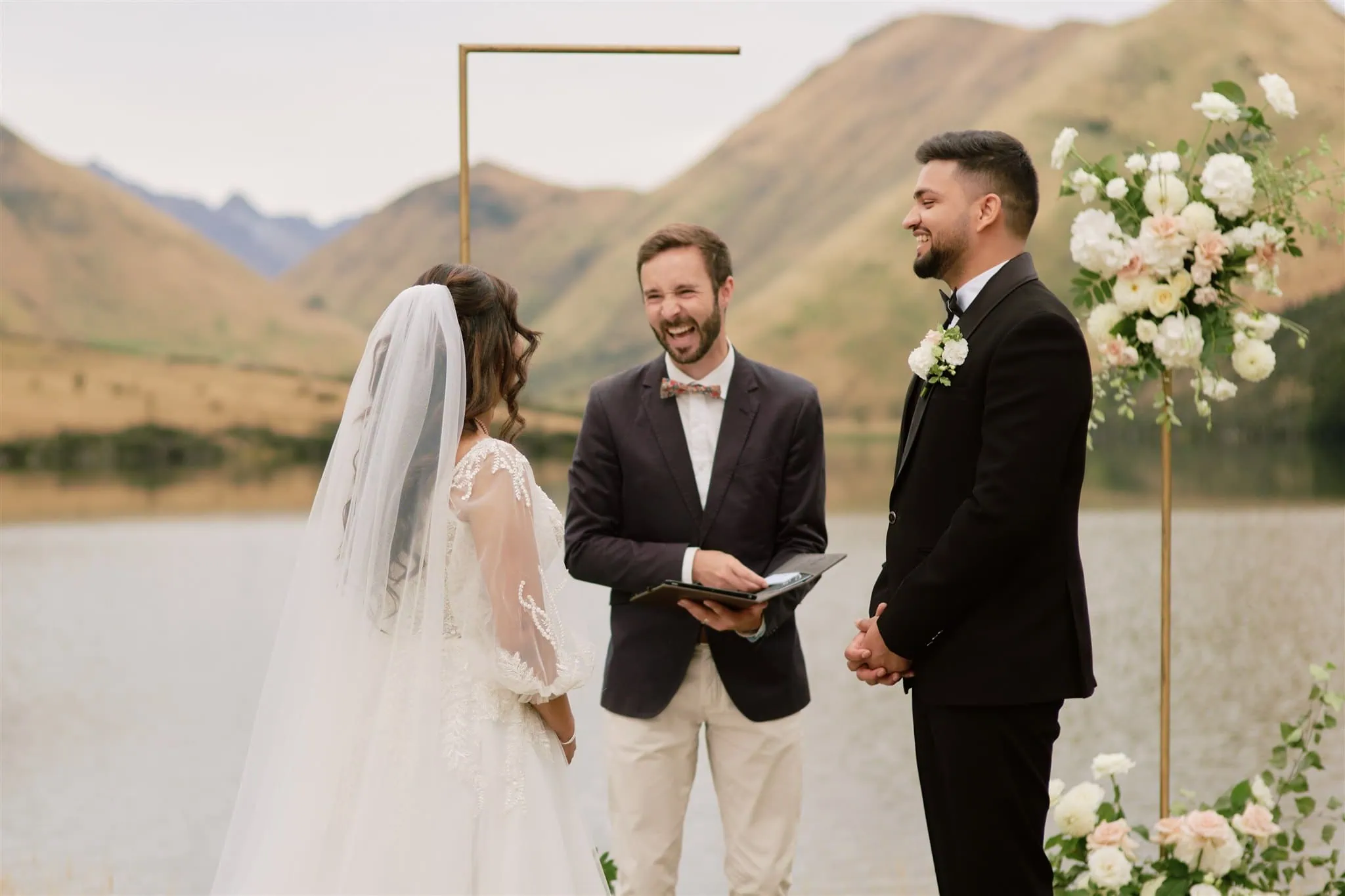queenstown new zealand heli wedding elopement photographer videographer | A Queenstown elopement ceremony by a lake, featuring Vidhya and Kartik exchanging vows with an officiant, framed by mountains and floral decorations.