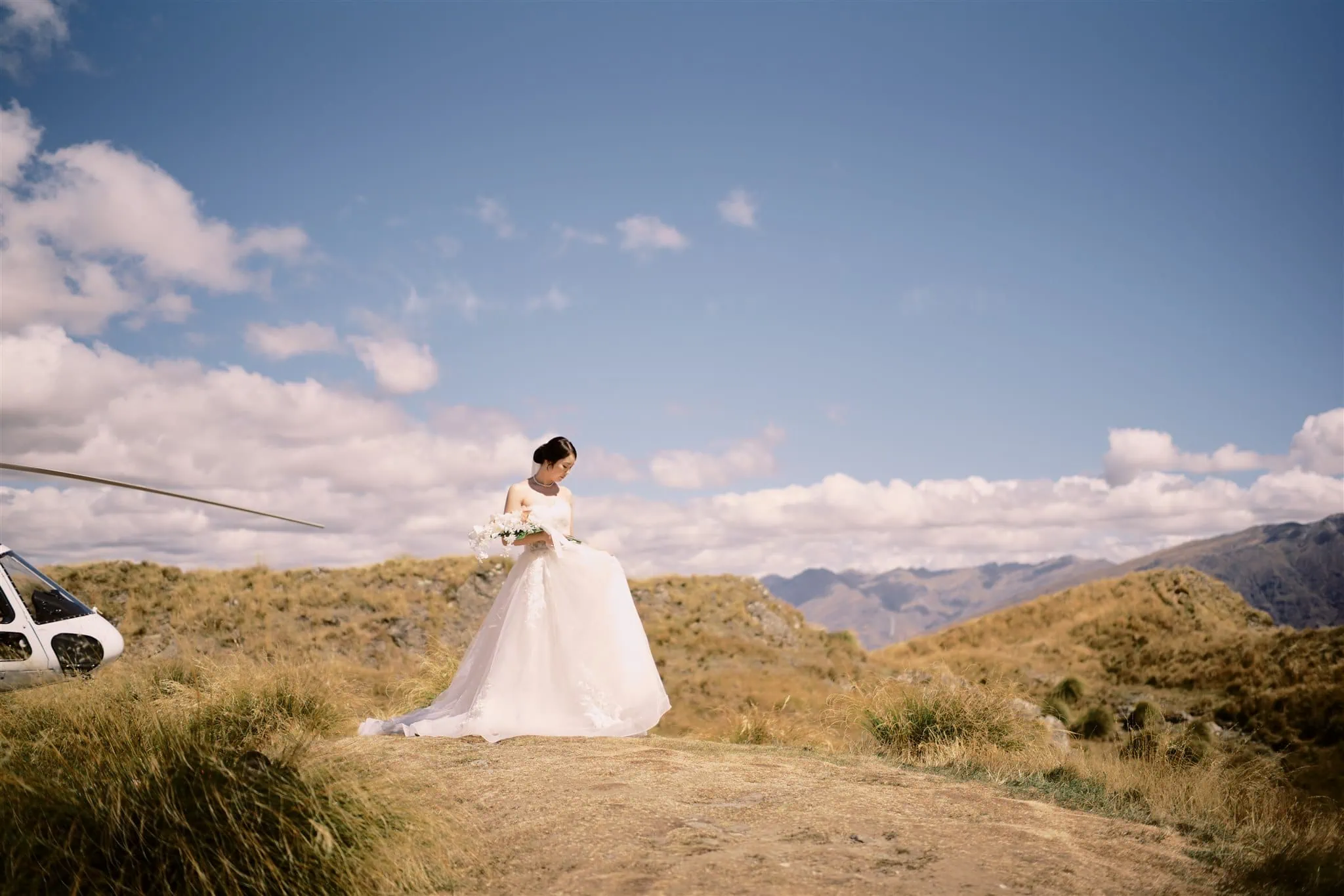 queenstown new zealand heli wedding elopement photographer videographer | A bride in a white dress holding a bouquet stands near a helicopter on a grassy hilltop, with mountains under a cloudy sky in the background, during her Queenstown elopement.