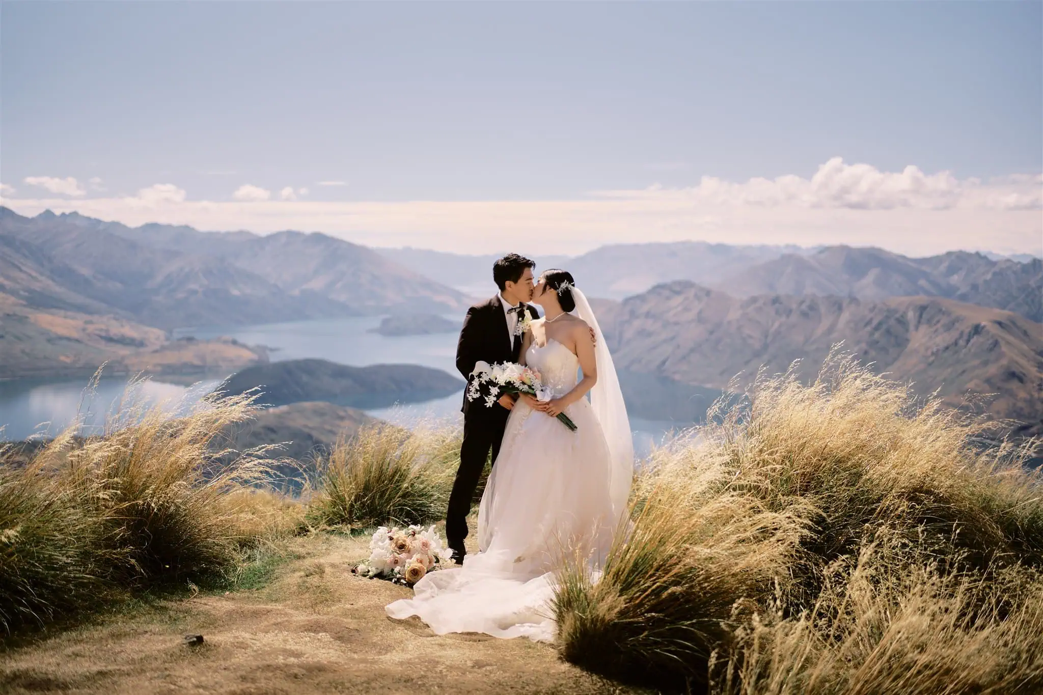 queenstown new zealand heli wedding elopement photographer videographer | Ryo and Tamano, a bride and groom, kissing atop a hill during their Queenstown elopement, overlooking a scenic lake surrounded by mountains, with tall grass in the foreground.