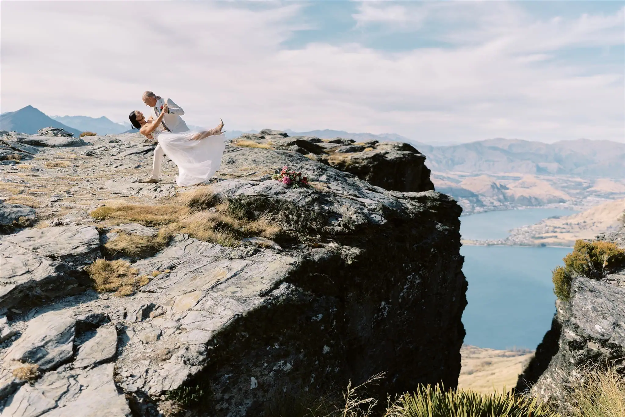 queenstown new zealand heli wedding elopement photographer videographer | A couple dressed in wedding attire share a dance on a rugged mountain cliff overlooking a lake and distant mountains during their Queenstown elopement.