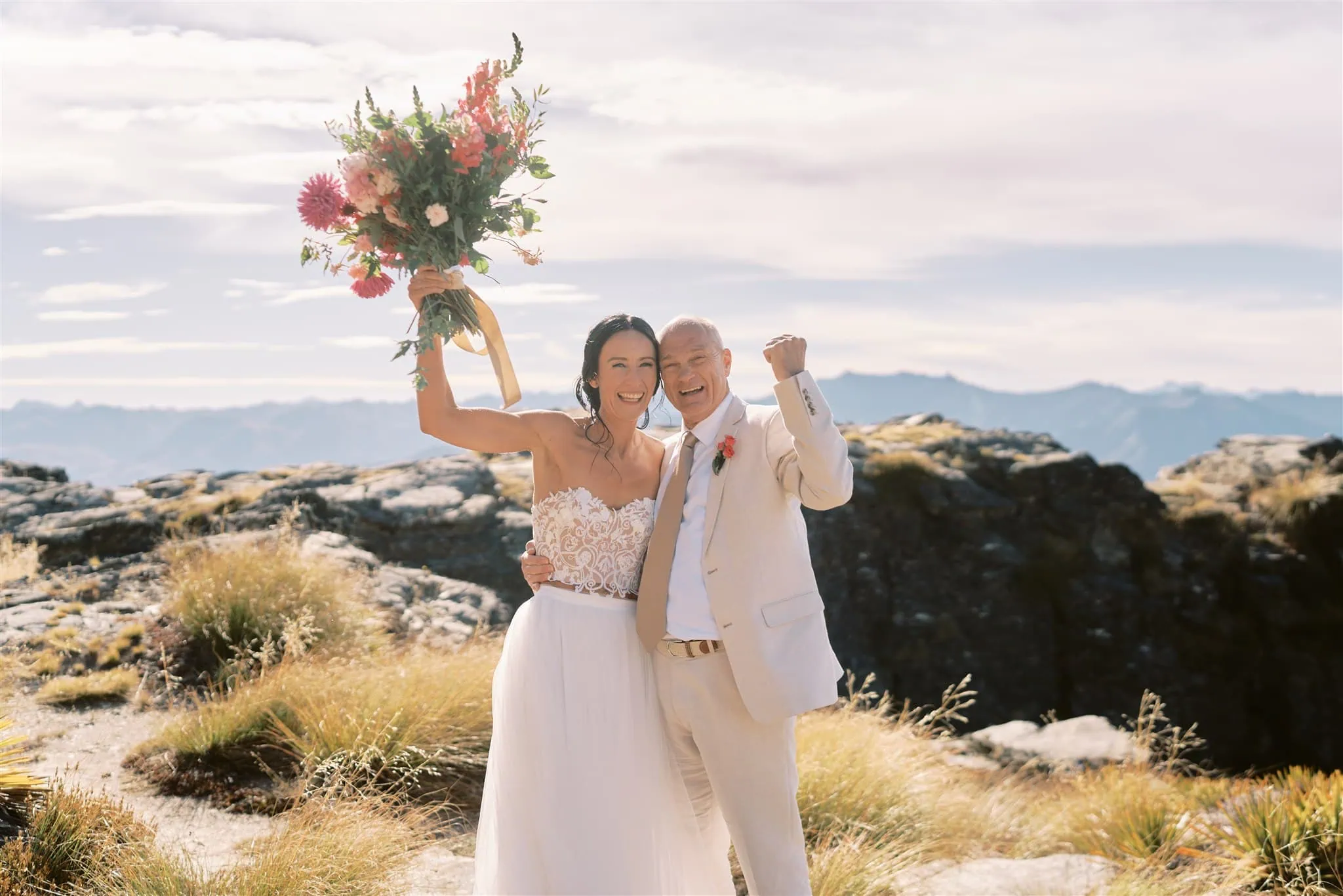 queenstown new zealand heli wedding elopement photographer videographer | Joyful bride and groom celebrating their Queenstown elopement on a mountain, the bride holding a vibrant bouquet, with scenic views in the background.