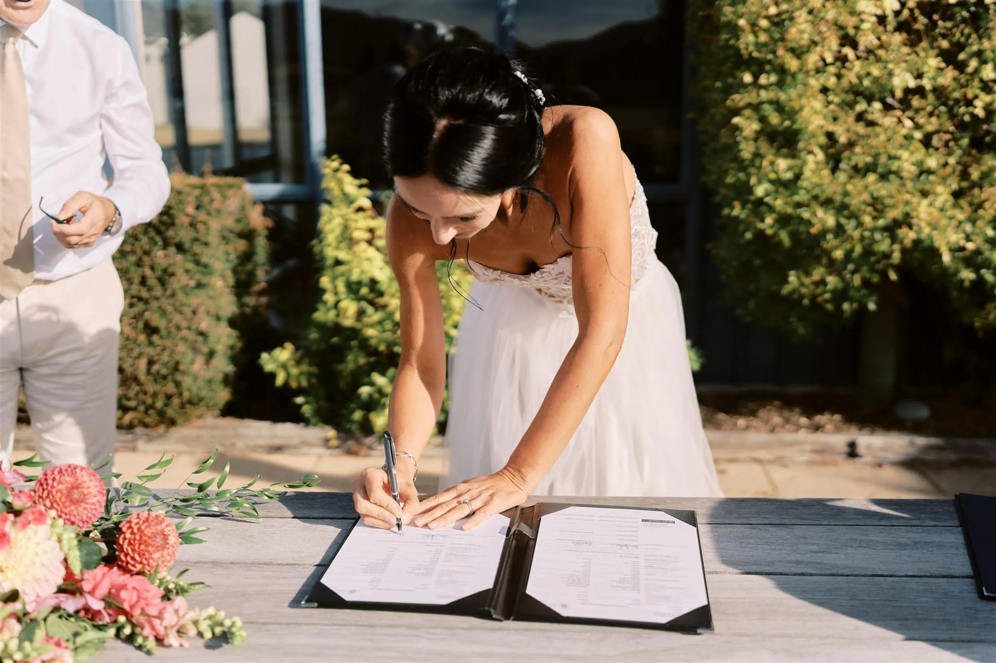 queenstown new zealand heli wedding elopement photographer videographer | A bride in a white dress signing her marriage certificate at a table, with a bouquet of colorful flowers nearby and a groom partially visible during their Queenstown elopement.