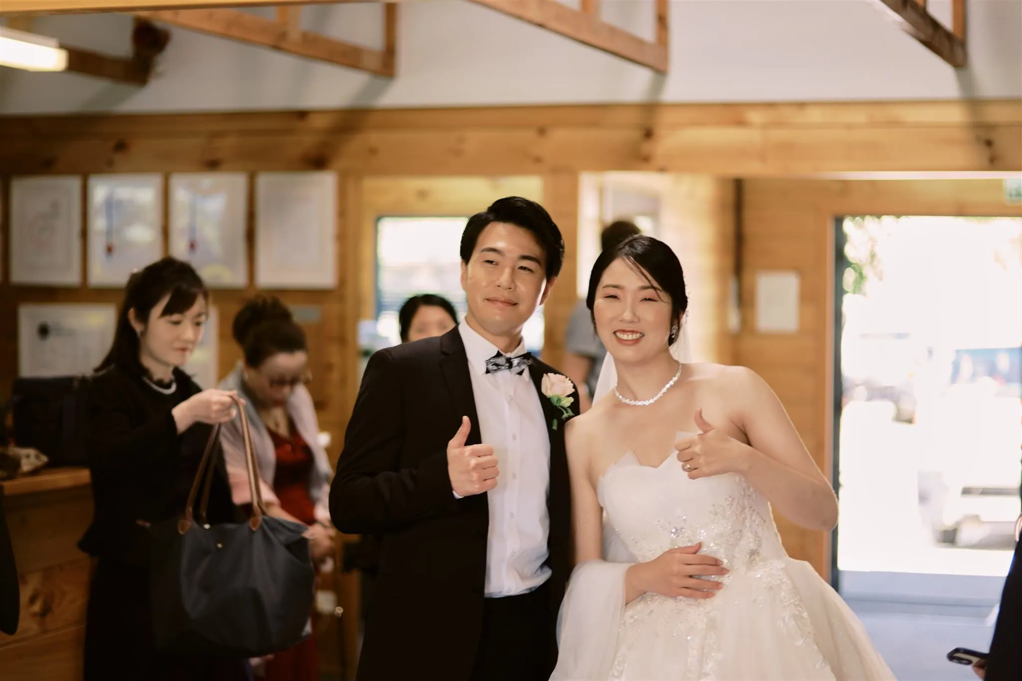 queenstown new zealand heli wedding elopement photographer videographer | A newlywed couple, Ryo & Tamano, posing with thumbs up in a festive indoor setting, surrounded by smiling guests.