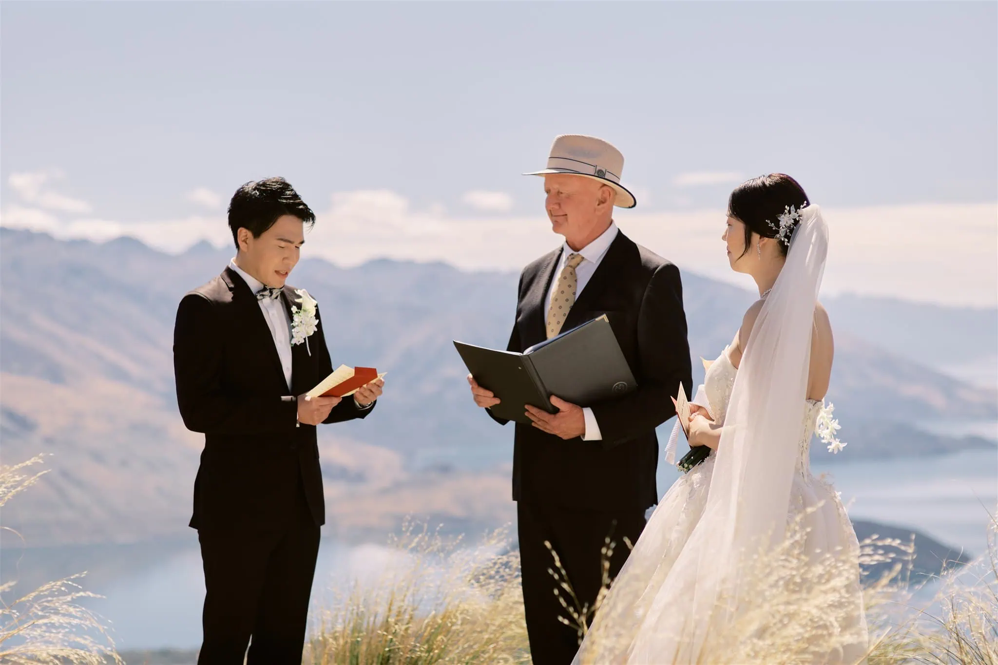 queenstown new zealand heli wedding elopement photographer videographer | An elopement ceremony outdoors with a groom, bride, and officiant standing in a field, mountains in the background.