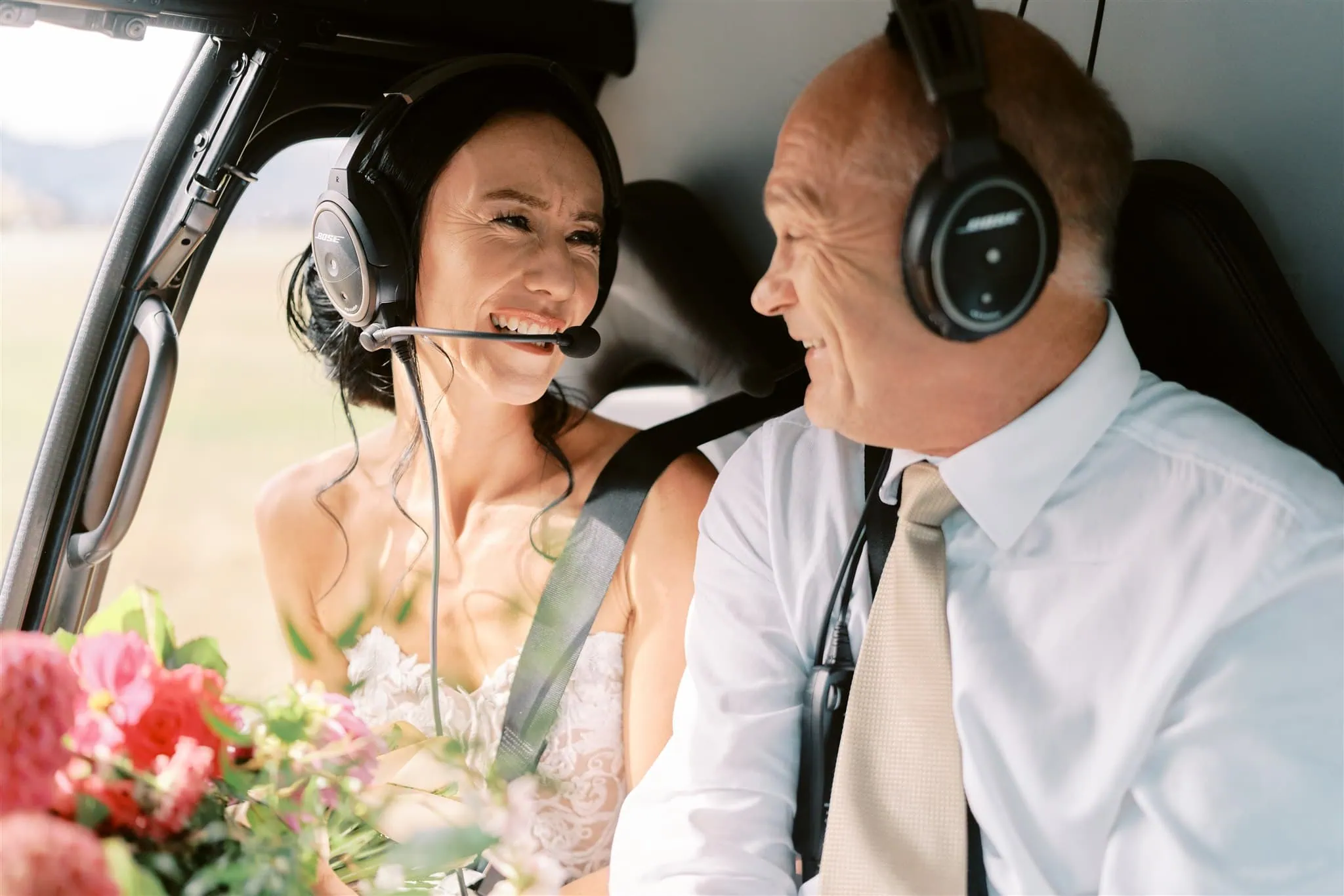 queenstown new zealand heli wedding elopement photographer videographer | Ian & Vivienne, a bride and groom wearing headsets, smile at each other inside a helicopter, surrounded by a bouquet of flowers.