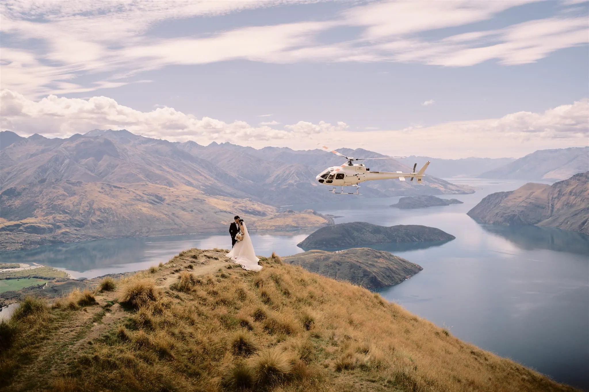 queenstown new zealand heli wedding elopement photographer videographer | A wedding couple stands on a hilltop near a lake in Queenstown with a helicopter flying overhead, surrounded by mountainous terrain.