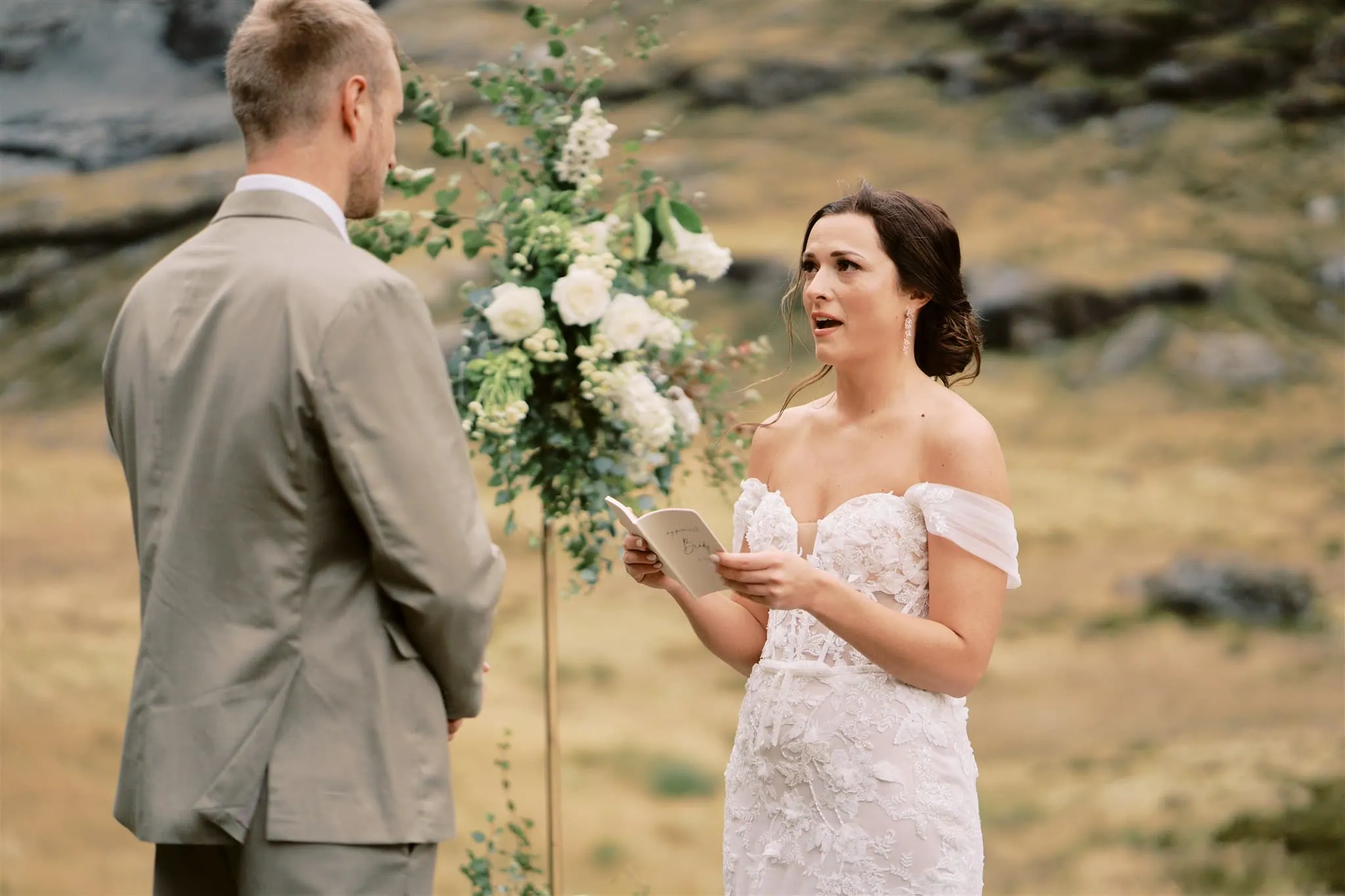 queenstown new zealand heli wedding elopement photographer videographer | A bride in a white dress reads her vows to the groom in an outdoor Queenstown elopement setting with a mountainous backdrop.