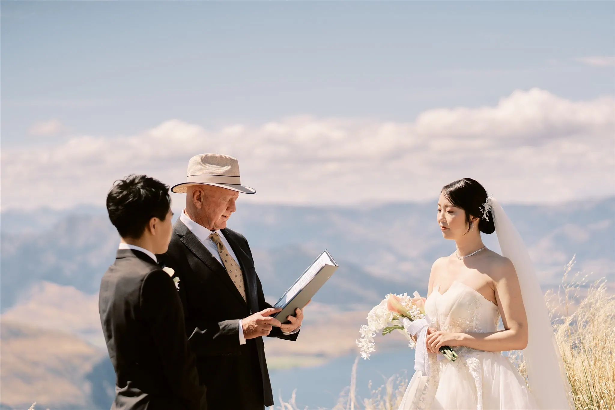 queenstown new zealand heli wedding elopement photographer videographer | Ryo and Tamano listen to an officiant during their Queenstown elopement ceremony, set against a scenic mountainous backdrop.