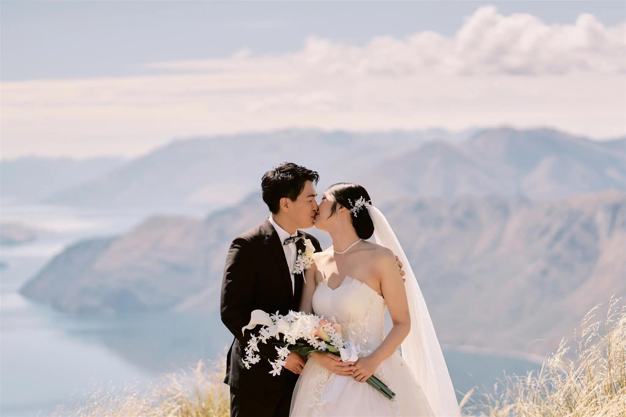 queenstown new zealand heli wedding elopement photographer videographer | Bride and groom kissing outdoors against a backdrop of mountains and a lake under a clear blue sky during their Queenstown elopement.
