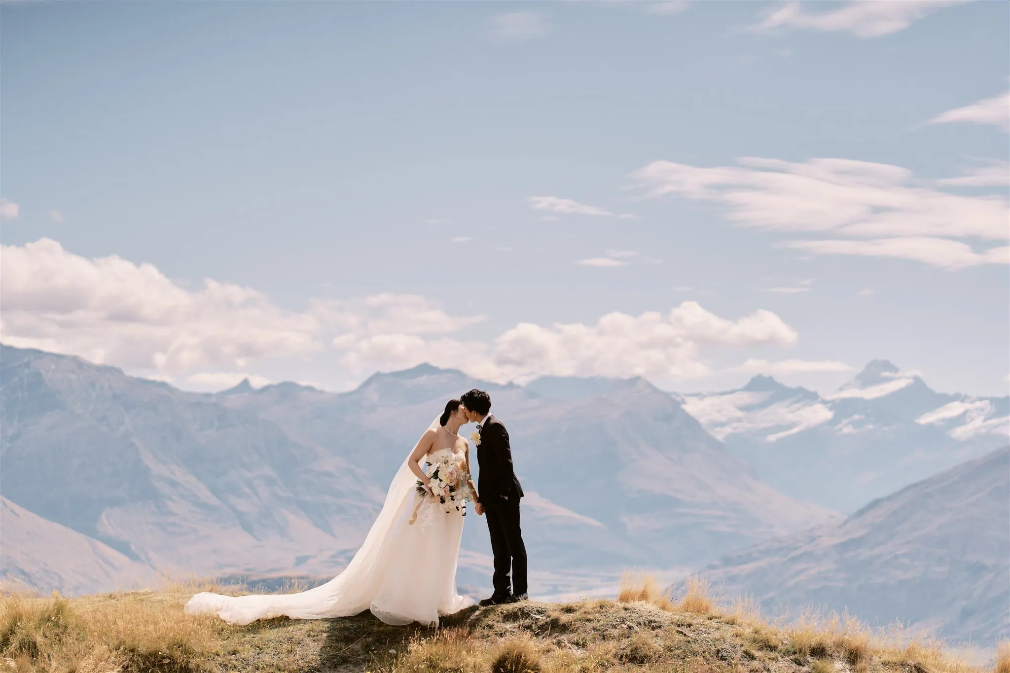 queenstown new zealand heli wedding elopement photographer videographer | A bride and groom kissing on a hilltop during their Queenstown elopement, with a sweeping view of distant mountains under a clear blue sky.