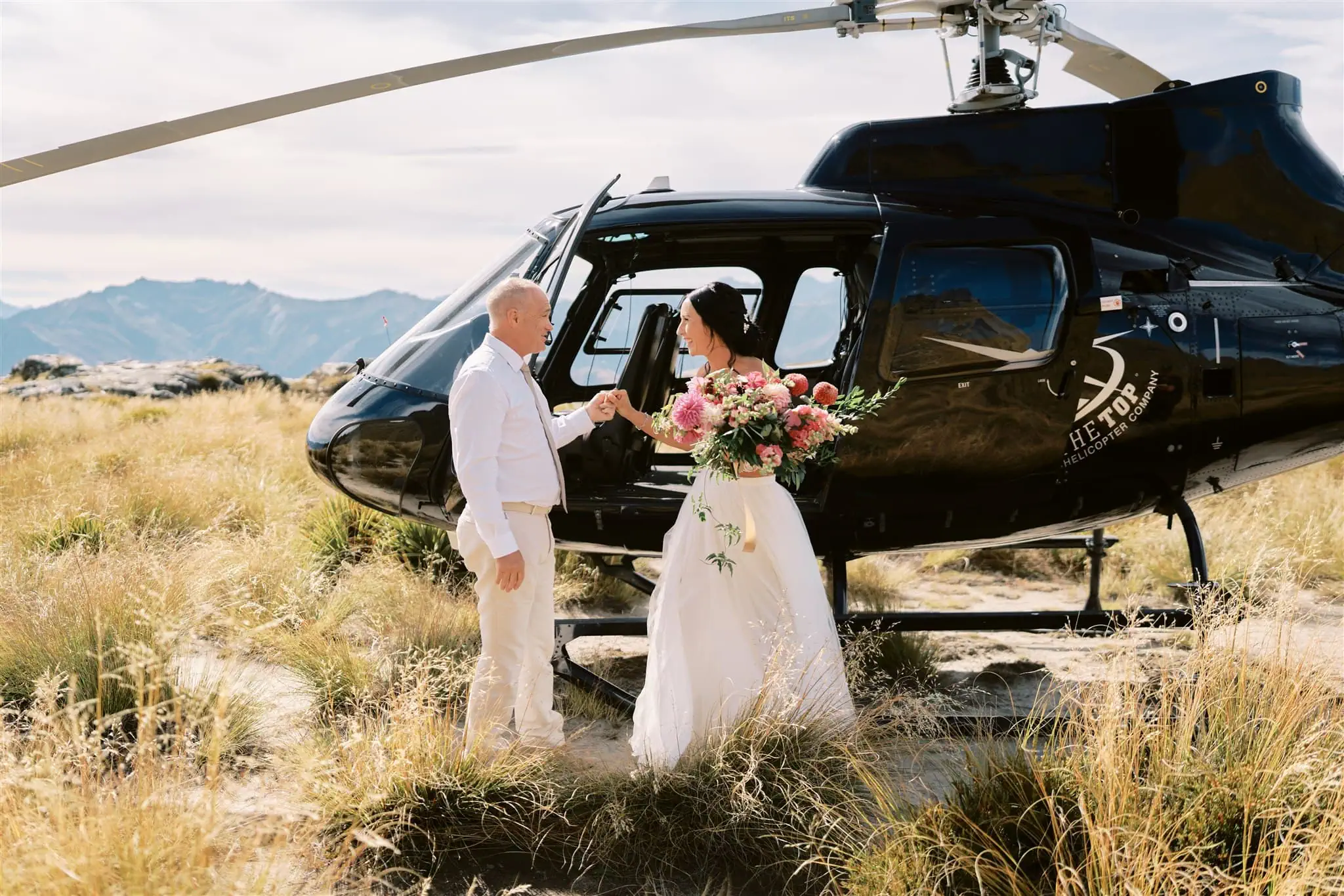 queenstown new zealand heli wedding elopement photographer videographer | Ian & Vivienne hold hands beside a black helicopter in a grassy field with mountains in the background.