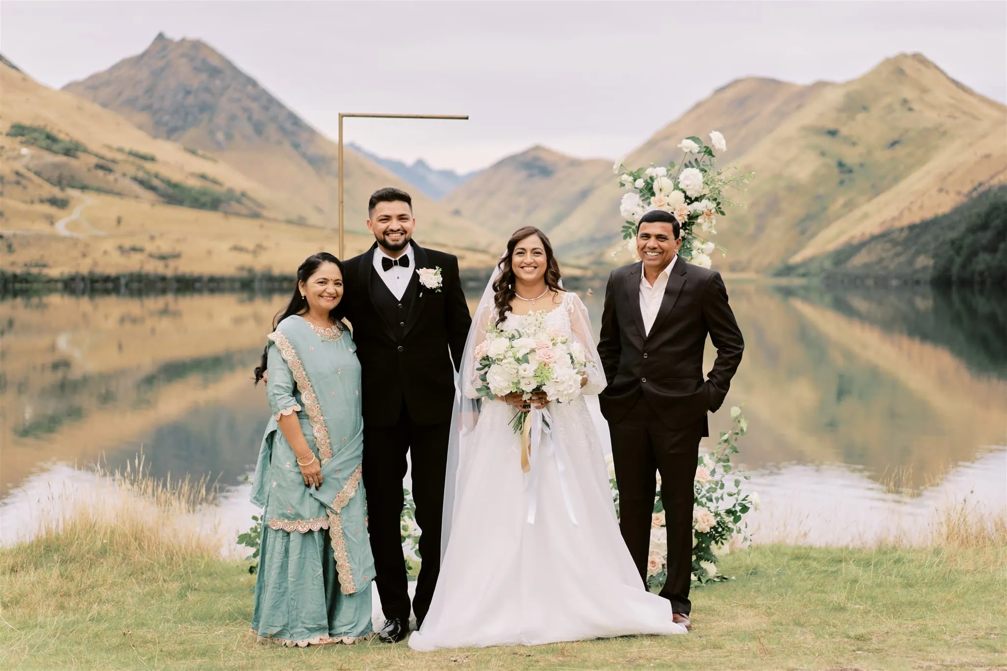 queenstown new zealand heli wedding elopement photographer videographer | A wedding party with two couples, including Vidhya & Kartik, stands by a serene lake in Queenstown, with mountains reflecting in the water and floral arrangements beside them.