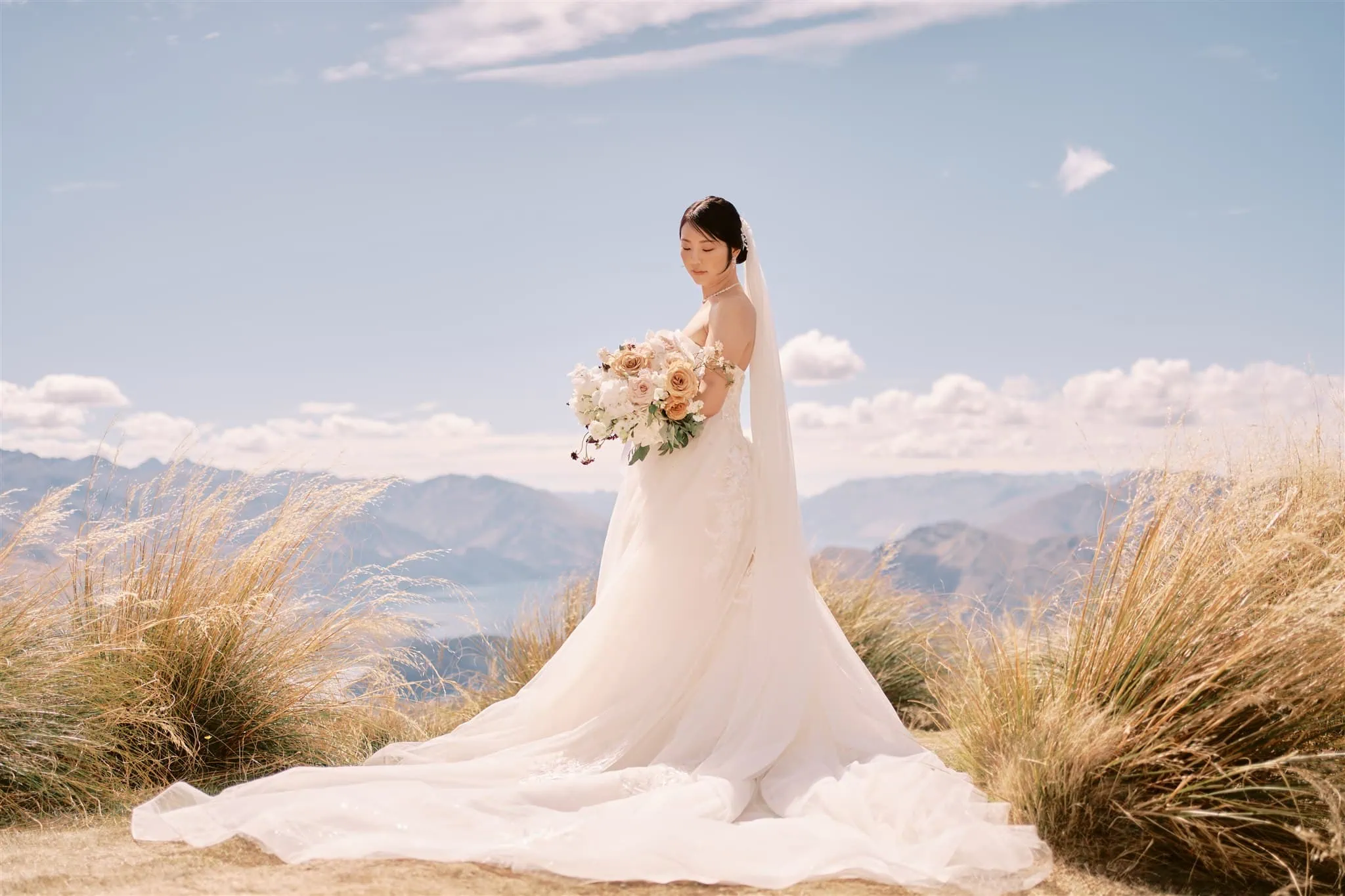 queenstown new zealand heli wedding elopement photographer videographer | A bride in a long white gown holding a bouquet stands amidst tall grass with a Queenstown Elopement backdrop under a clear sky.