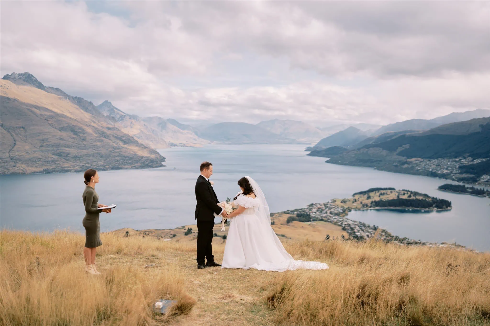 queenstown new zealand heli wedding elopement photographer videographer | A couple in wedding attire stands at an outdoor ceremony, perfectly captured by Queenstown Wedding Photography, with a picturesque lake and mountain backdrop, accompanied by an officiant.