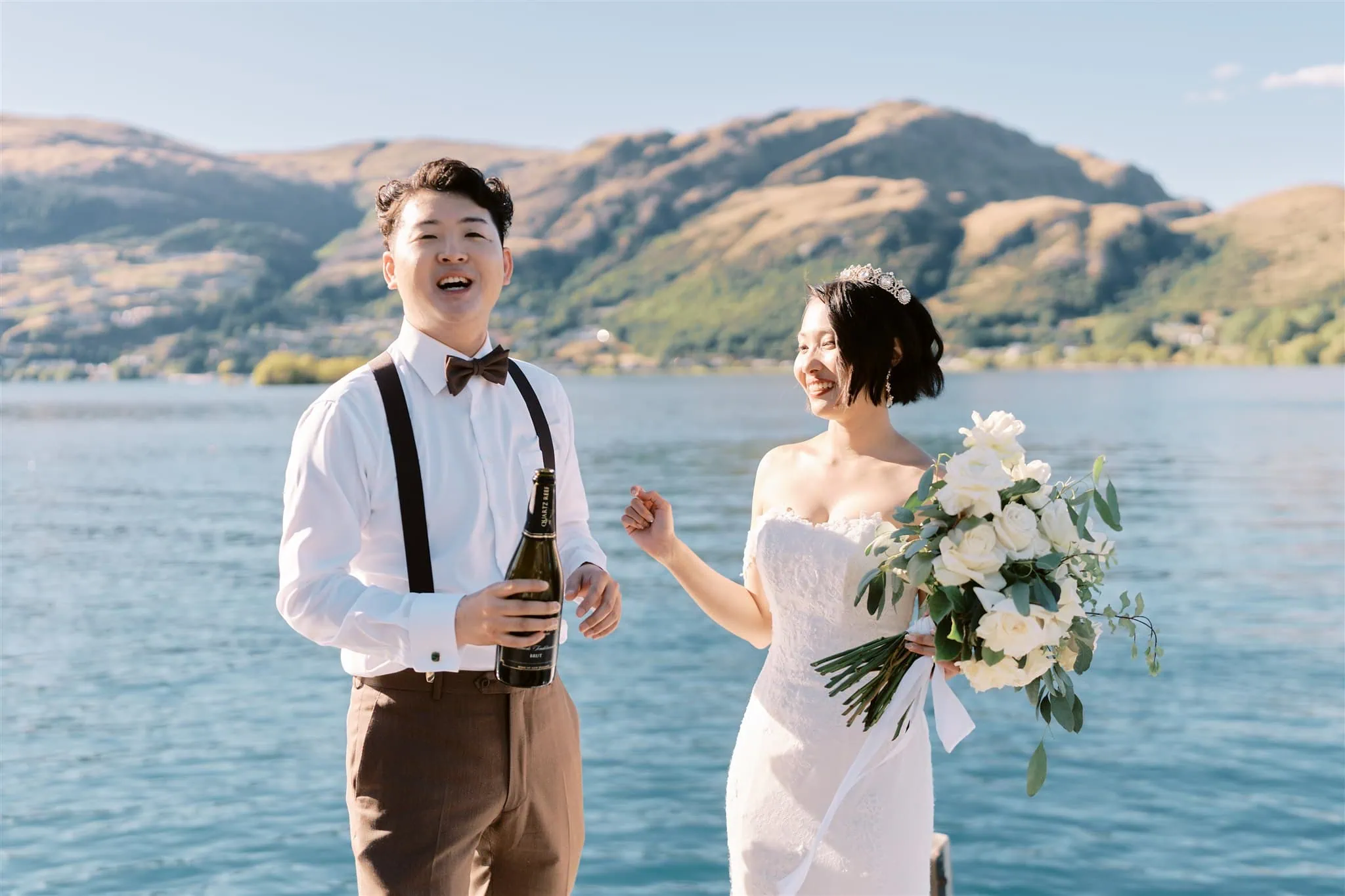queenstown new zealand heli wedding elopement photographer videographer | A joyful bride and groom holding champagne and flowers by a lake with mountains in the background on a sunny day during their Queenstown Heli Elopement.