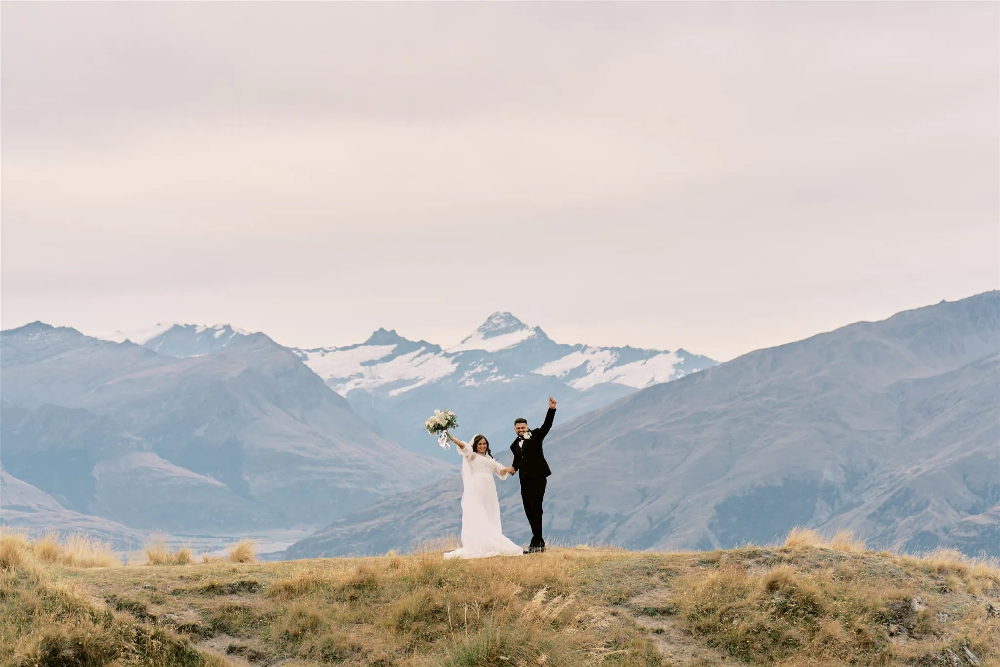 queenstown new zealand heli wedding elopement photographer videographer | A newlywed couple celebrates with raised arms on a grassy hill, mountains in the background under a soft sky during their Queenstown elopement.
