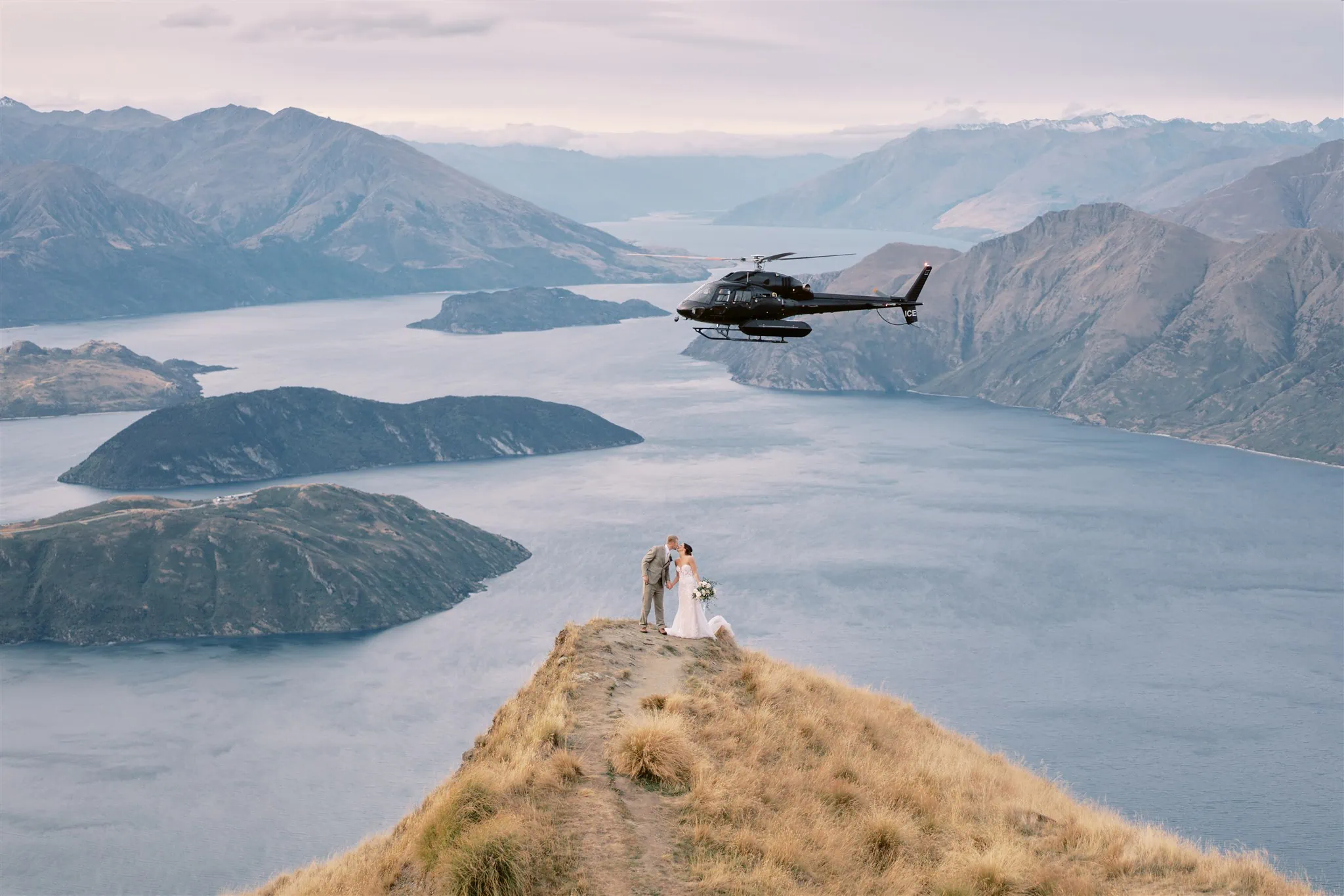 queenstown new zealand heli wedding elopement photographer videographer | A bride and groom standing on a narrow ridge overlooking a scenic lake with mountains in Queenstown, as a helicopter flies nearby.