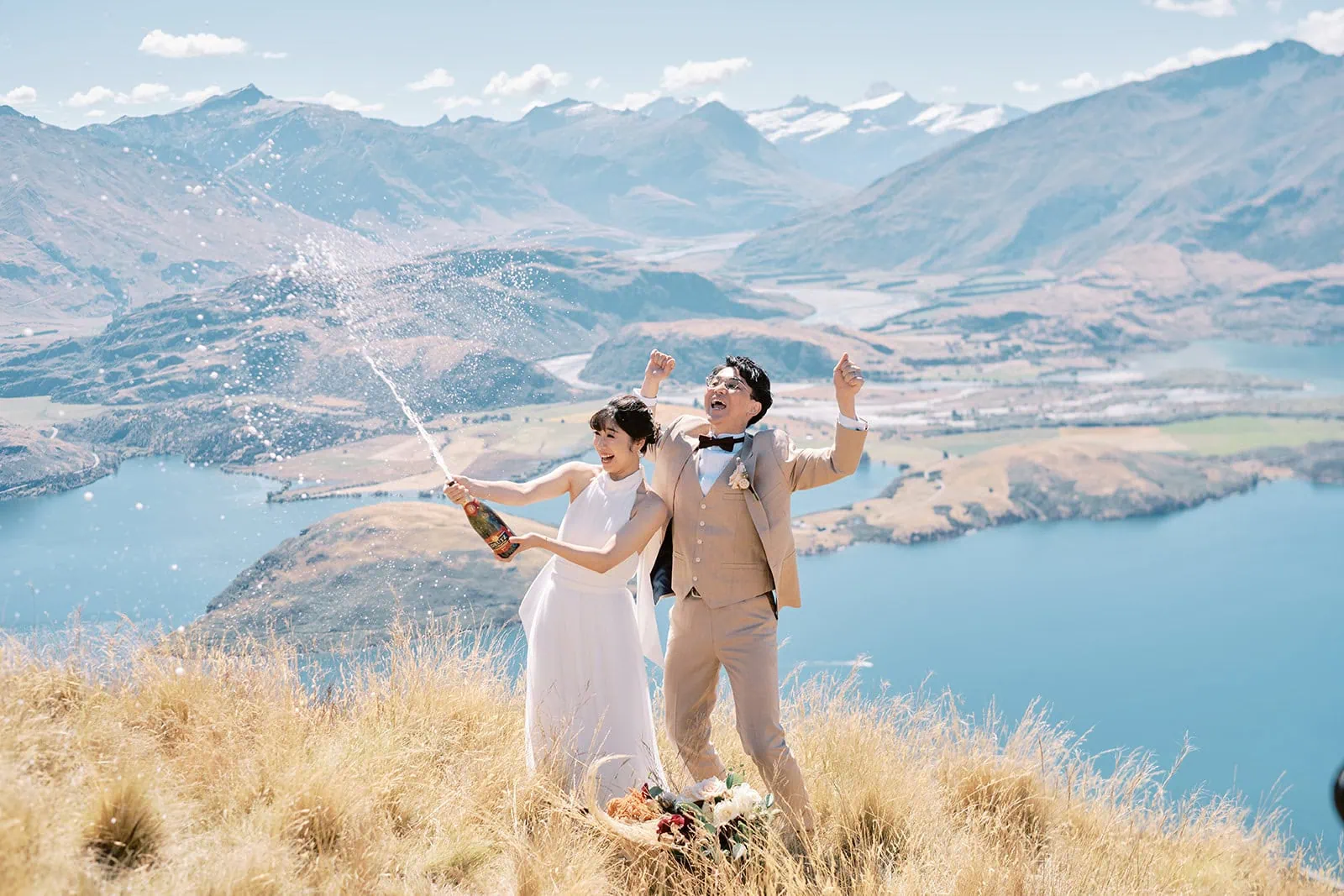 queenstown new zealand heli wedding elopement photographer videographer | A couple enjoying a Queenstown pre-wedding shoot with champagne, overlooking a scenic lake and mountains on a sunny day.