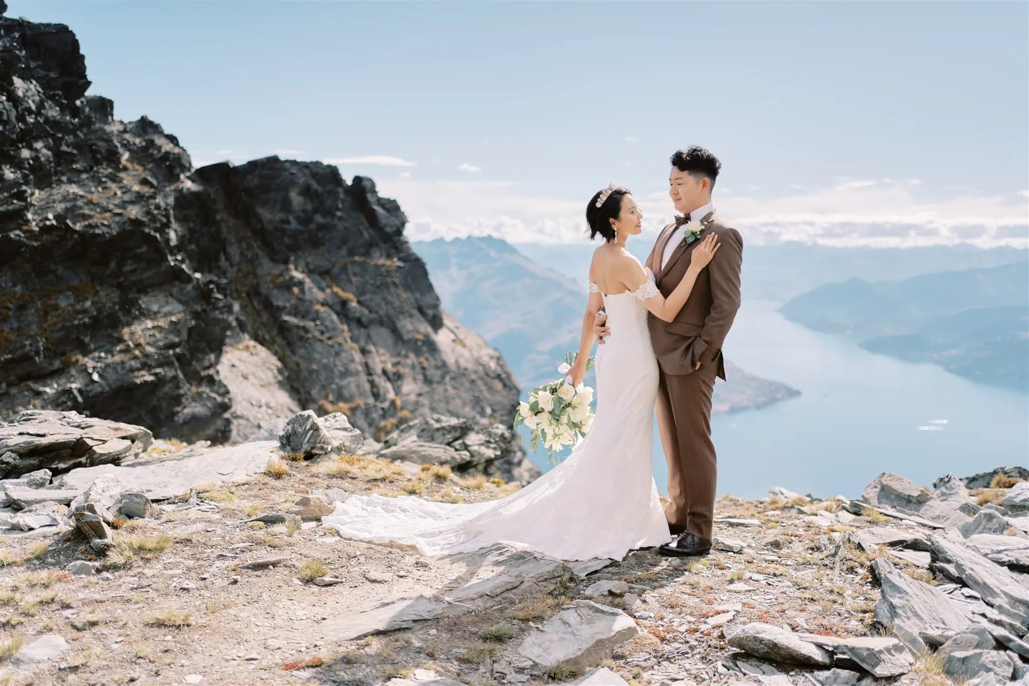 queenstown new zealand heli wedding elopement photographer videographer | A couple in wedding attire embracing on a mountain overlooking a lake under a clear blue sky during their Queenstown Heli Elopement.