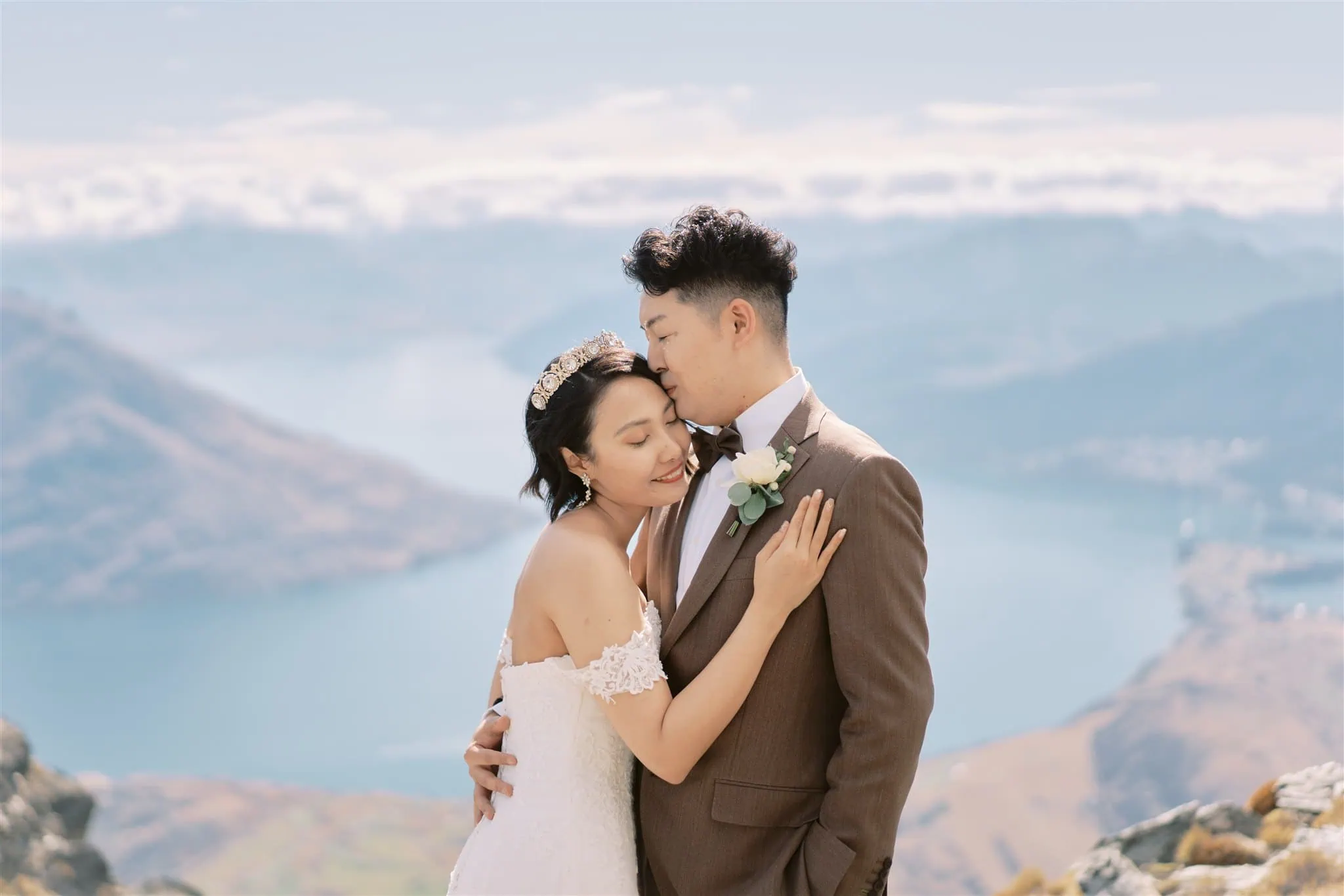 queenstown new zealand heli wedding elopement photographer videographer | Azusa and Yuhei, a bride and groom embracing on a mountain, with a serene lake and hills in the background under a clear blue sky.