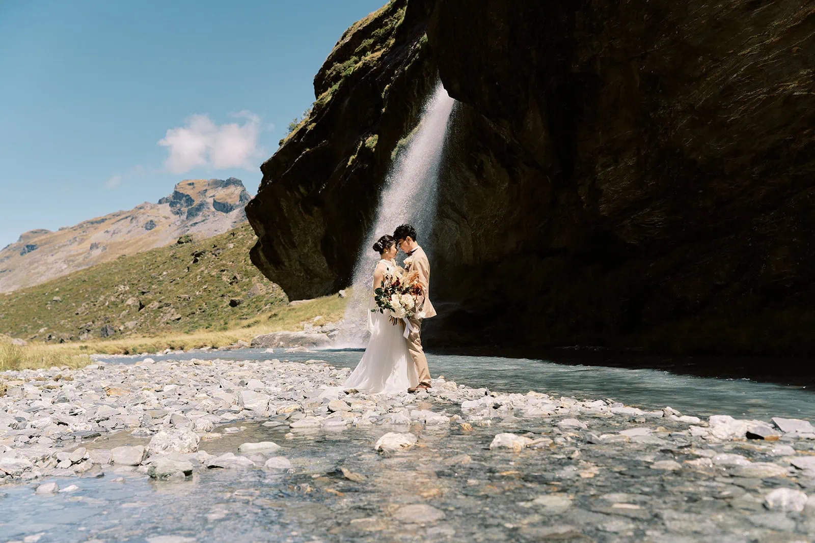 queenstown new zealand heli wedding elopement photographer videographer | Nozomi and Ippei in wedding attire embracing near a waterfall with rocky terrain under a clear blue sky.