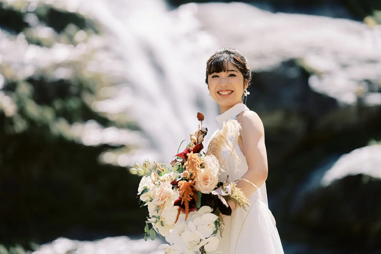 queenstown new zealand heli wedding elopement photographer videographer | A bride holding a bouquet smiles brightly in front of a waterfall on a sunny day during her Queenstown pre-wedding shoot.