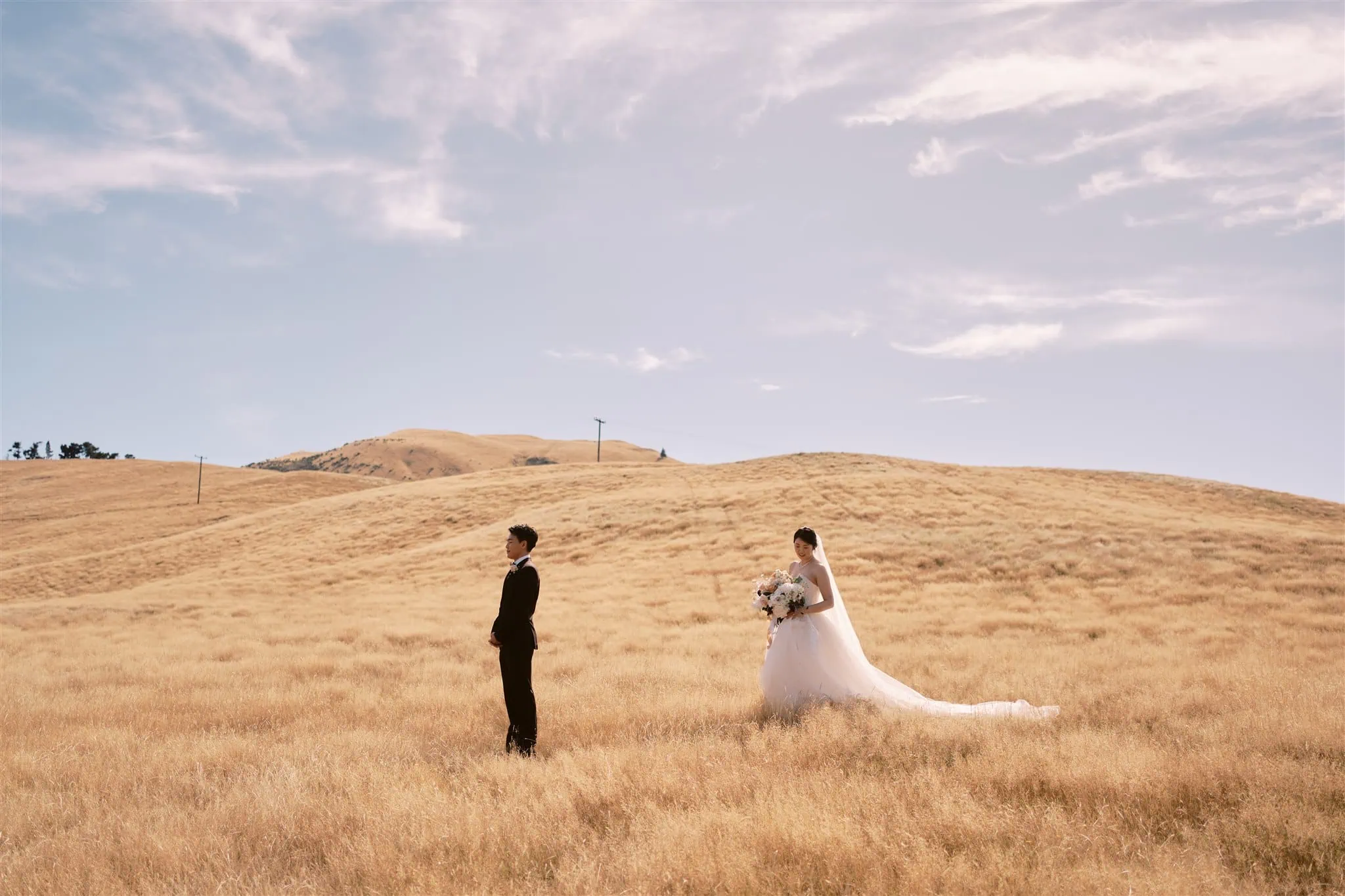 queenstown new zealand heli wedding elopement photographer videographer | A bride and groom standing apart in a vast field of golden grass under a clear sky, with rolling hills in the background during their Queenstown elopement.