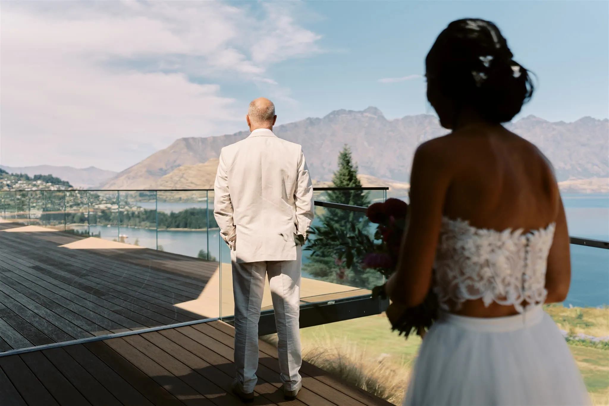queenstown new zealand heli wedding elopement photographer videographer | Bride in focus on the right watching the groom, who faces a scenic lake and mountains from a balcony during their Queenstown elopement.