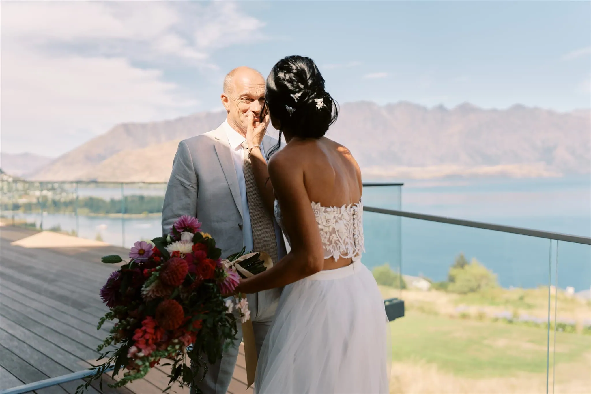 queenstown new zealand heli wedding elopement photographer videographer | Ian & Vivienne, eloping in Queenstown, share a tender moment on a balcony overlooking a scenic lake and mountains. Ian touches Vivienne's face gently as they look into each other's eyes.