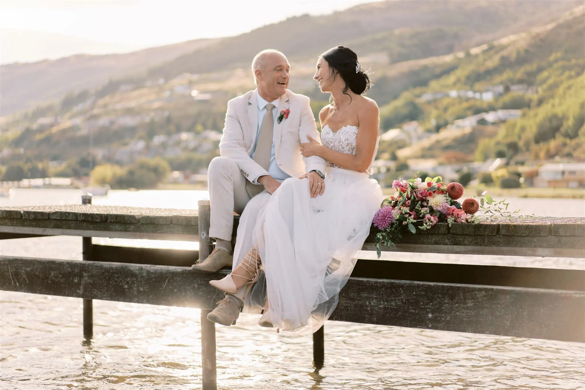 queenstown new zealand heli wedding elopement photographer videographer | Ian, an elderly groom, and Vivienne, a young bride, smiling at each other while sitting on a wooden beam by a lake during sunset in Queenstown.
