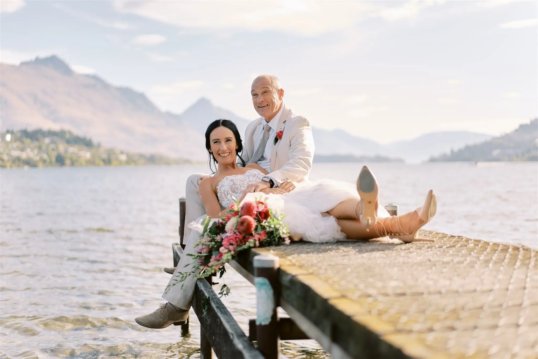 queenstown new zealand heli wedding elopement photographer videographer | Newlywed couple Ian & Vivienne sitting and smiling on a lakeside dock, with mountains in the background on a sunny day.