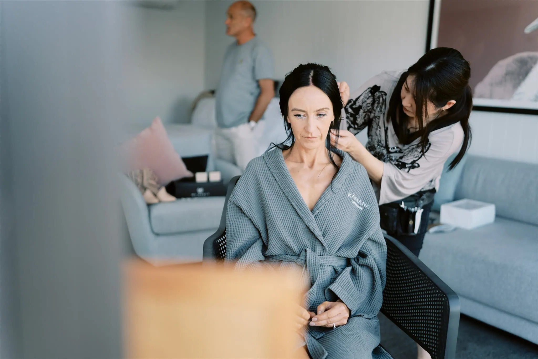 queenstown new zealand heli wedding elopement photographer videographer | A woman in a grey robe getting her hair styled by another woman in a modern living room, with a man in the background, during a Queenstown elopement.