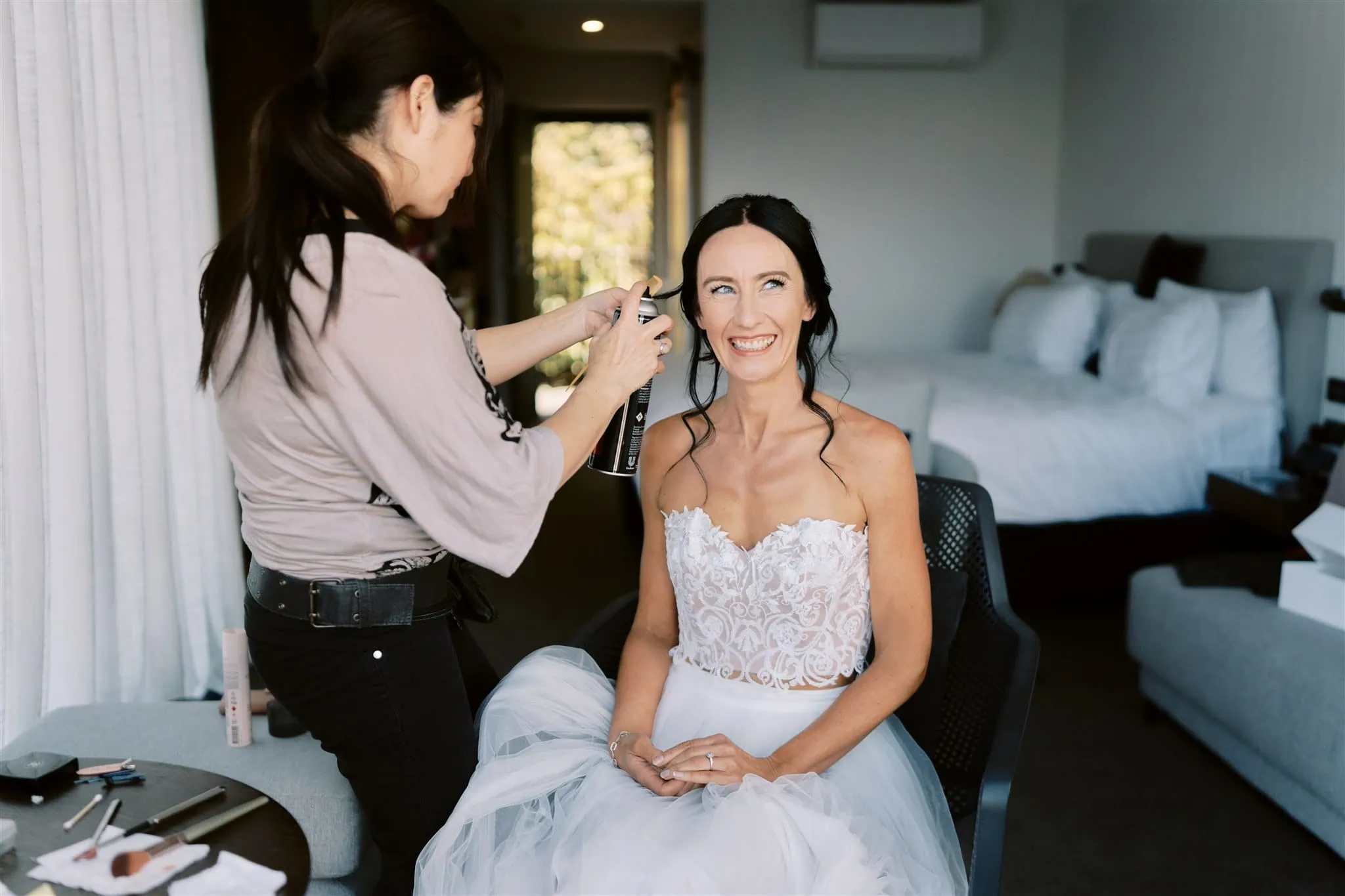 queenstown new zealand heli wedding elopement photographer videographer | A bride in a white dress smiles while a stylist sprays her hair in a hotel room during her Queenstown elopement.
