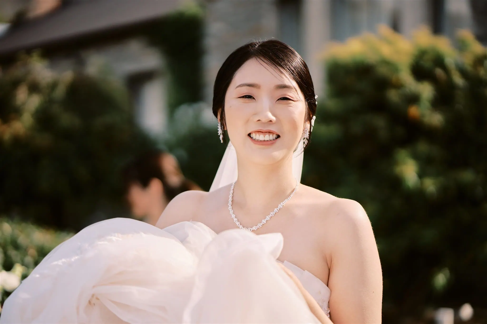queenstown new zealand heli wedding elopement photographer videographer | A bride in a white gown smiling joyfully, carrying her dress, with a soft focus background of a garden and a building during her Queenstown elopement.