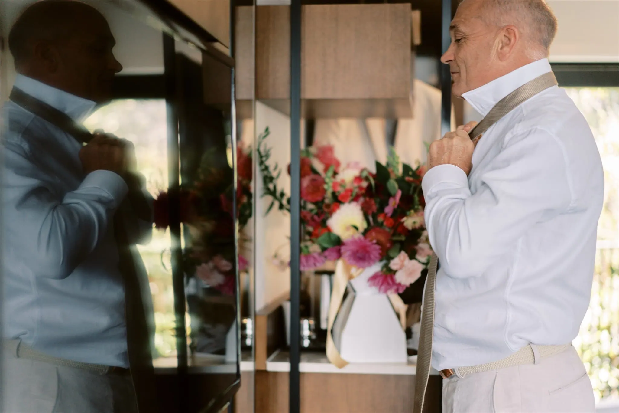 queenstown new zealand heli wedding elopement photographer videographer | An older man adjusting his tie while looking at his reflection in a wardrobe mirror, with a vase of flowers nearby, prepared for Ian & Vivienne's Queenstown elopement.