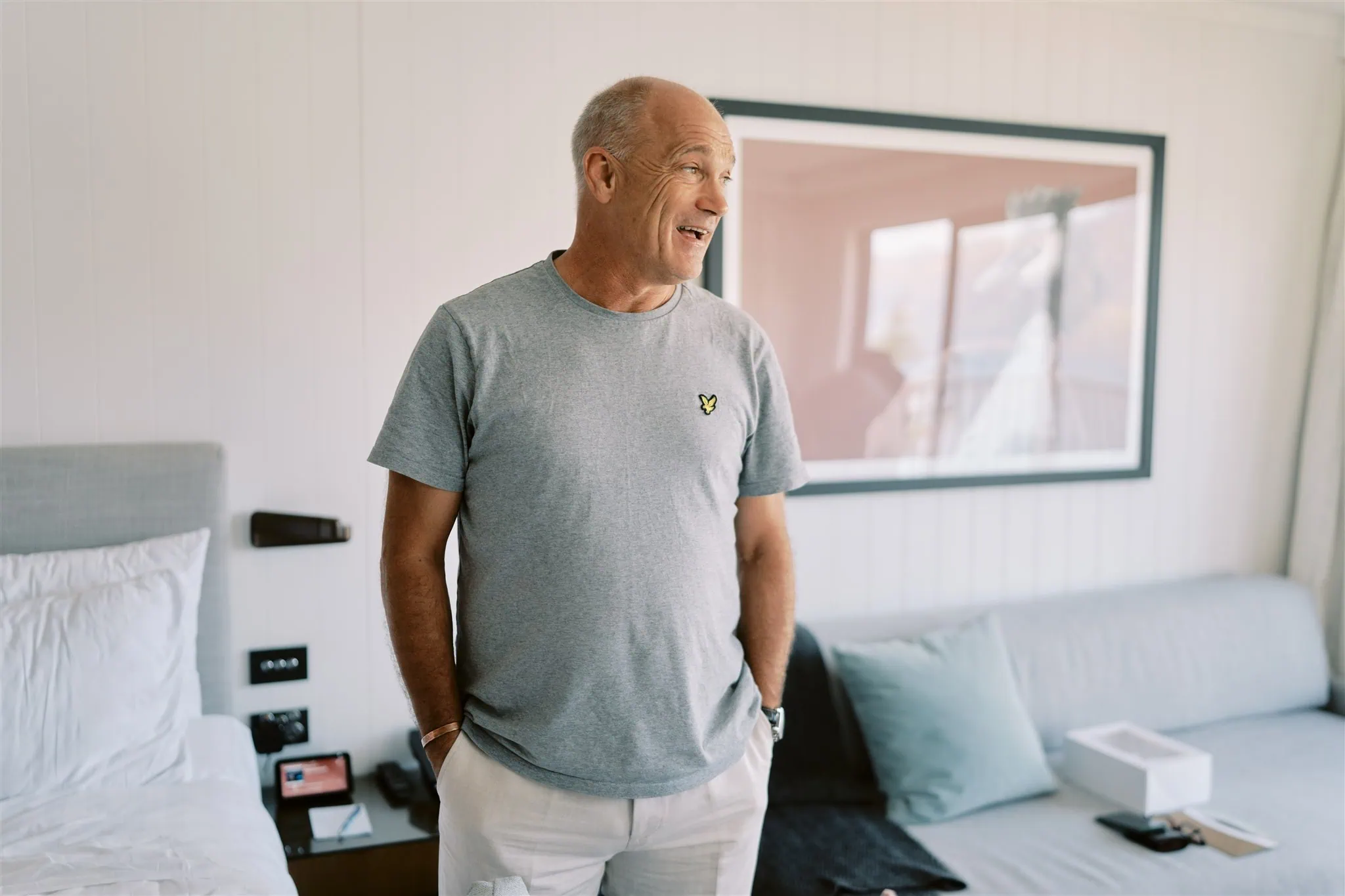 queenstown new zealand heli wedding elopement photographer videographer | A smiling older man standing in a bedroom, wearing a gray t-shirt and white pants, with a framed picture and a bed in the background, captured during an elopement photography session.