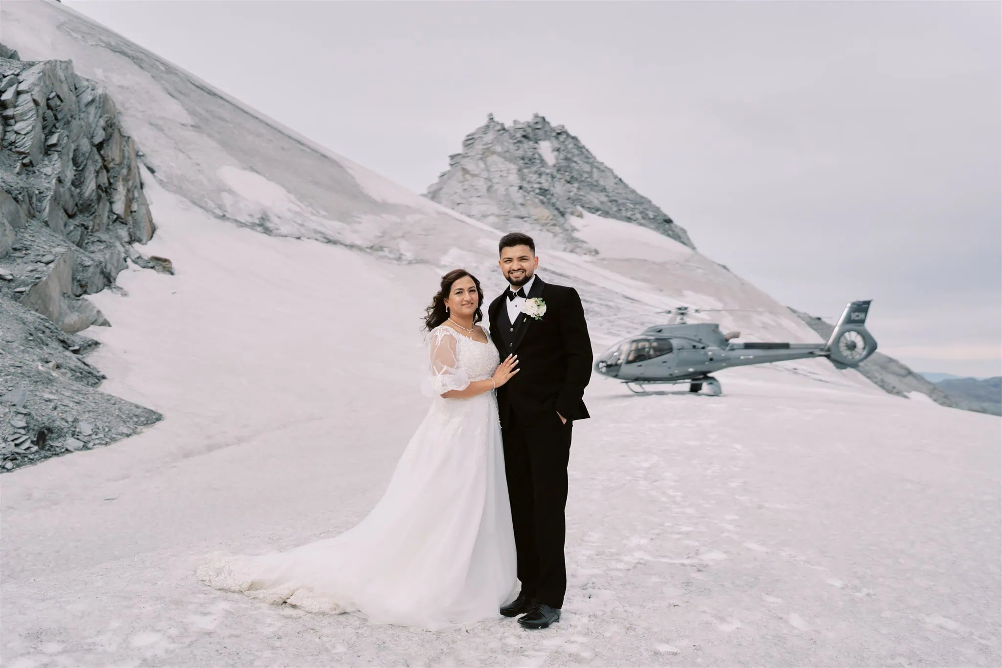 queenstown new zealand heli wedding elopement photographer videographer | VidhyA and Kartik pose on a snowy mountain landscape with a helicopter parked in the background during their Queenstown elopement.