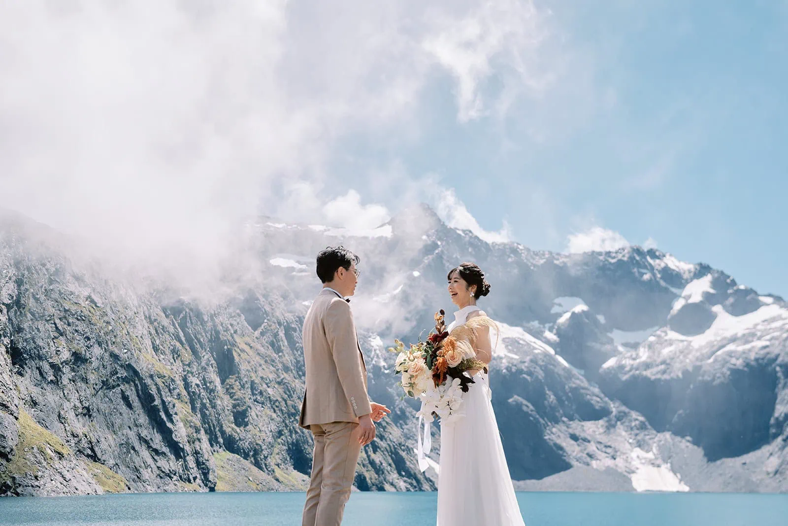 queenstown new zealand heli wedding elopement photographer videographer | A newlywed couple holding hands by a mountain lake in Queenstown, the bride holding a bouquet, with snowy peaks and blue sky in the background.