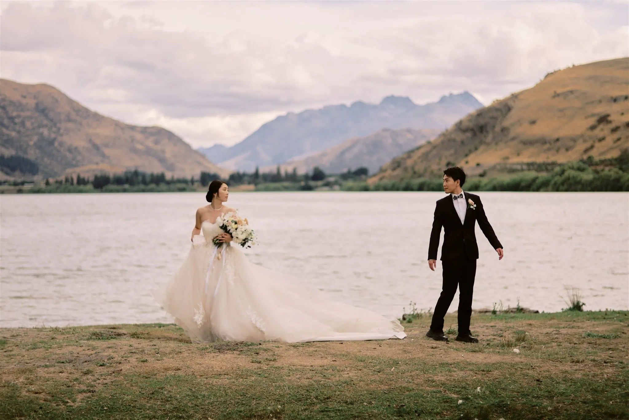 queenstown new zealand heli wedding elopement photographer videographer | A bride in a flowing gown and a groom in a tuxedo walking apart on a Queenstown lakeside path, with mountains in the background.