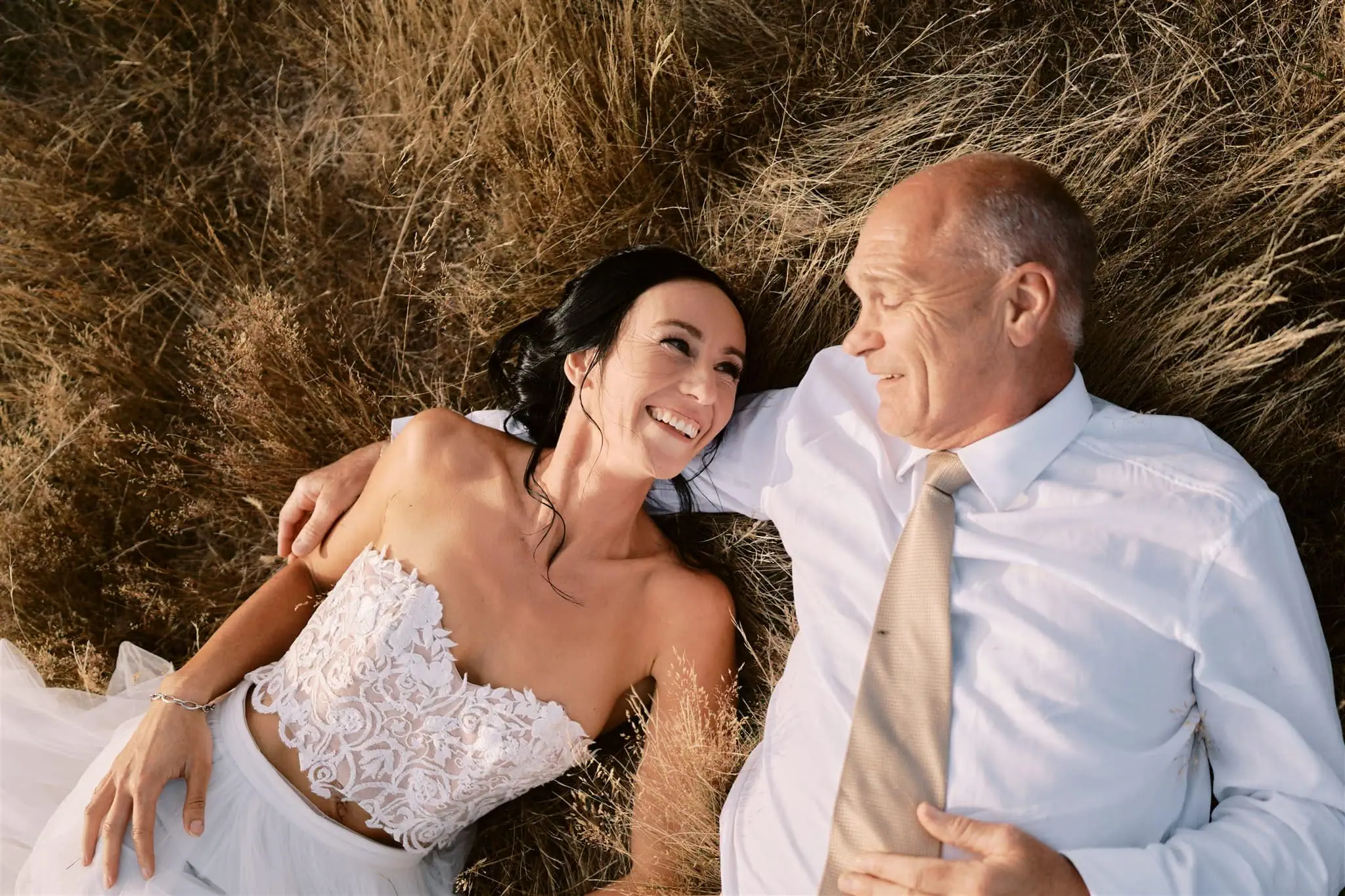 queenstown new zealand heli wedding elopement photographer videographer | A bride in a white dress and a groom in a suit lie in a field during their Queenstown elopement, smiling at each other.