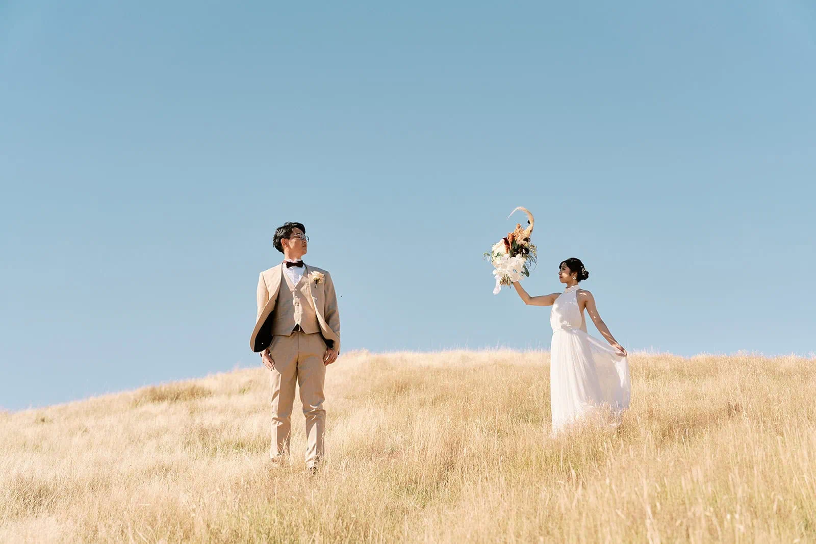 queenstown new zealand heli wedding elopement photographer videographer | Nozomi and Ippei walking on a grassy hill under a clear blue sky, with Nozomi tossing a flower bouquet.