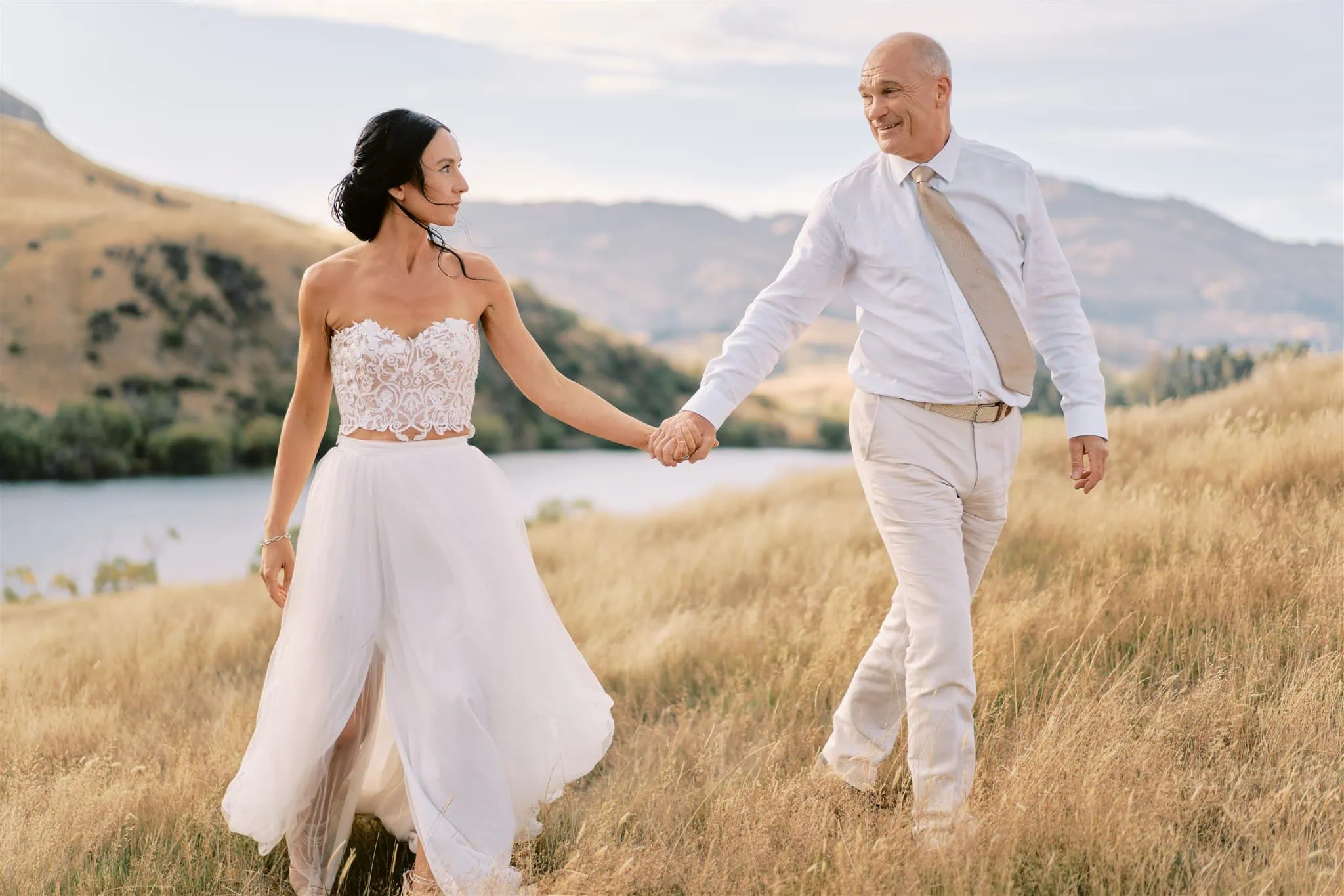 queenstown new zealand heli wedding elopement photographer videographer | A bride in a white dress and a groom in a white shirt holding hands and walking through a grassy field near a lake during their Queenstown elopement.