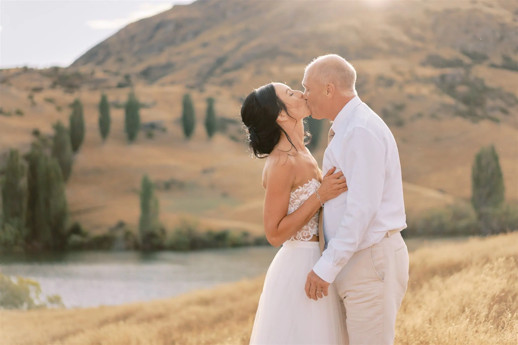 queenstown new zealand heli wedding elopement photographer videographer | A couple kisses in a scenic Queenstown landscape with a tranquil river and golden hills bathed in soft sunlight.