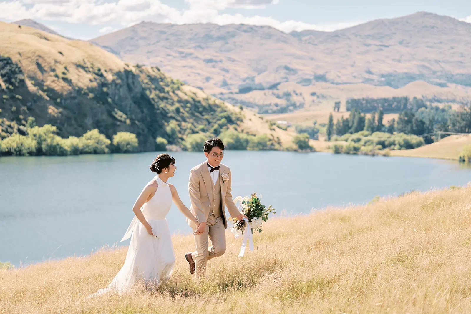 queenstown new zealand heli wedding elopement photographer videographer | A newlywed couple joyfully walking hand in hand through a golden field near a lake, with mountains in the background on a sunny day during their Queenstown Pre-Wedding Shoot.